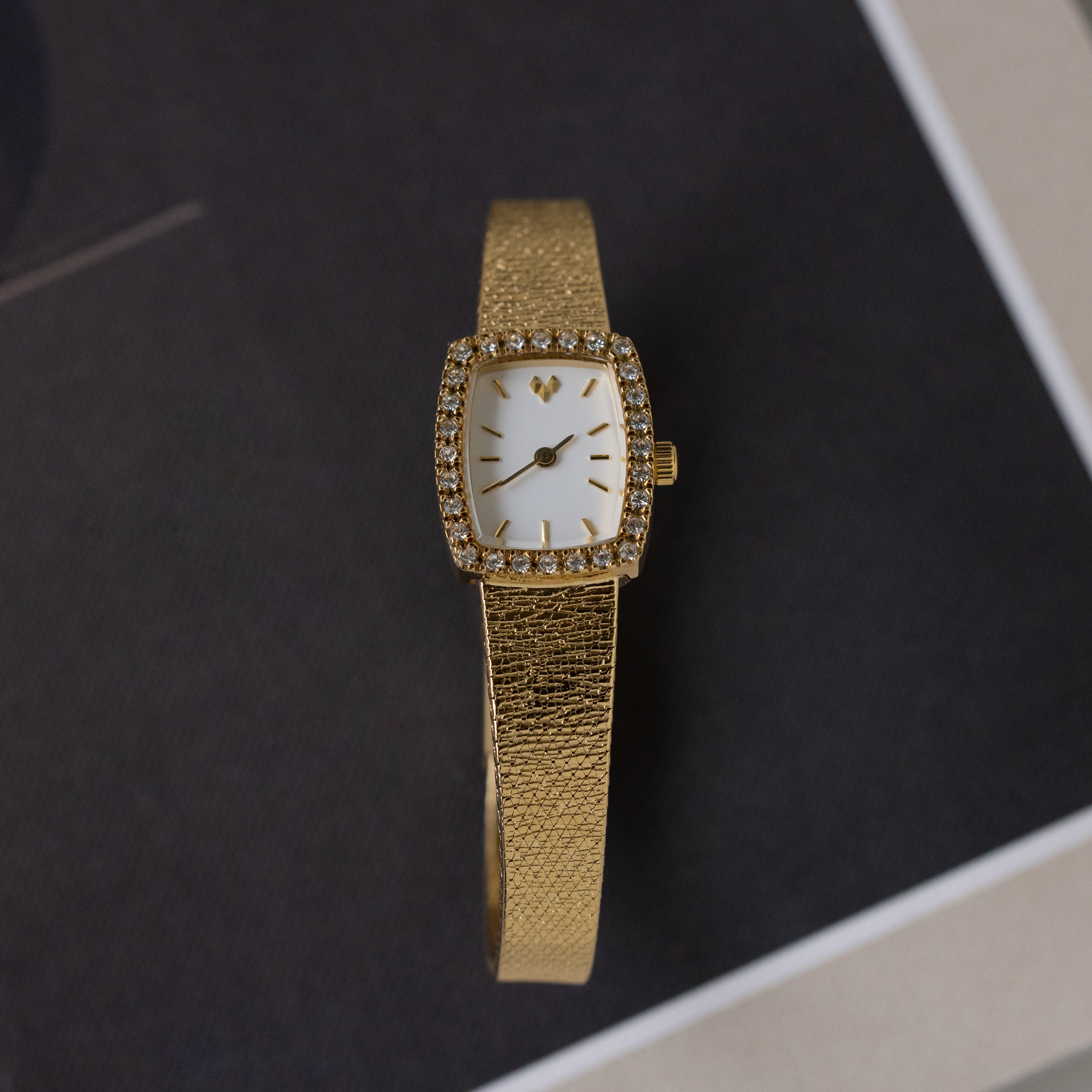 The Petite Mesh Timepiece features a gold rectangular face with diamond-like stones around the dial, shown elegantly on a dark surface.