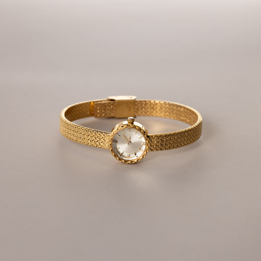 Gold round braided watch with a textured band displayed flat on a neutral surface.

