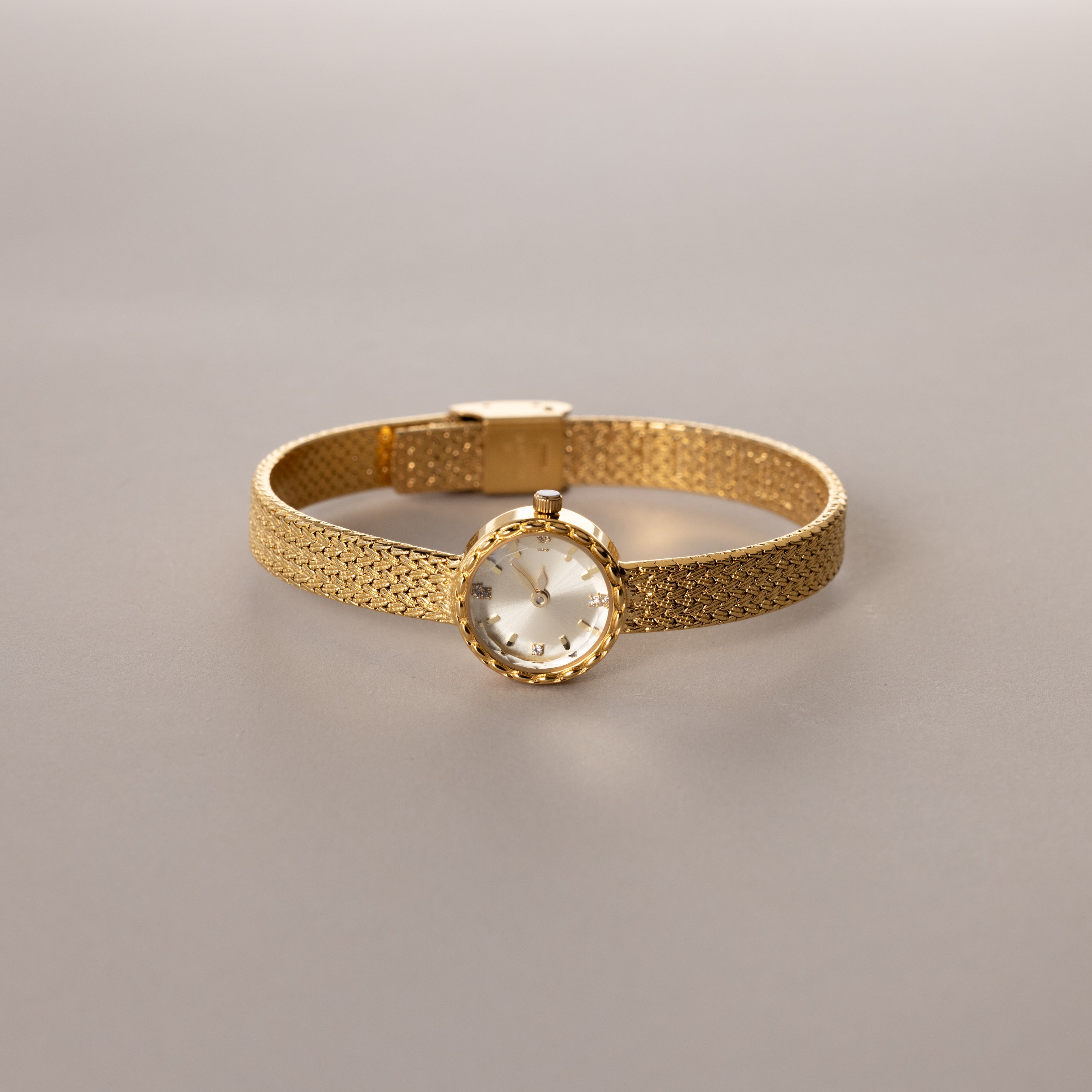 Gold round braided watch with a textured band displayed flat on a neutral surface.

