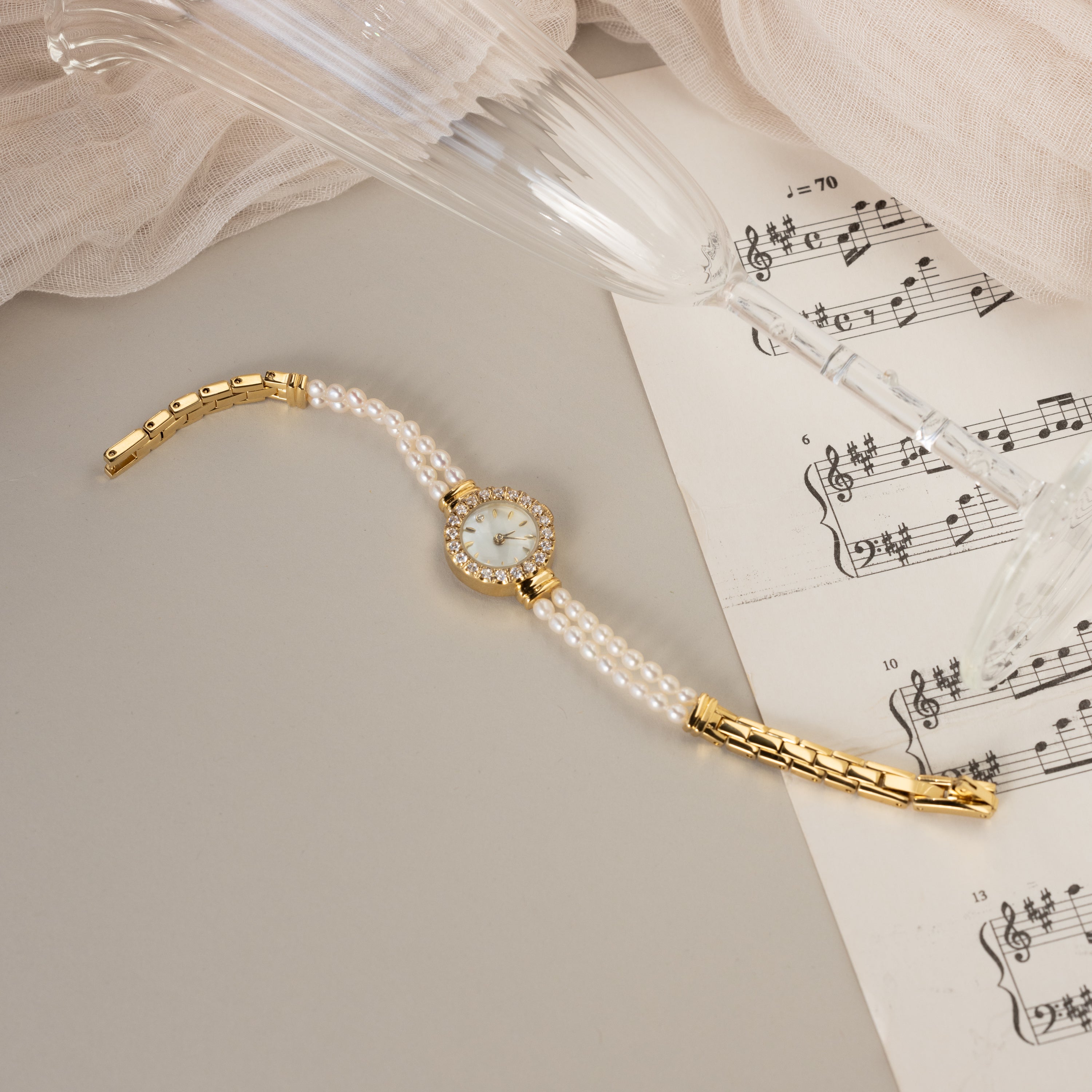 The Pearl Link Timepiece with diamond accents sits on sheet music next to a tilted champagne glass and beige fabric, radiating vintage elegance.