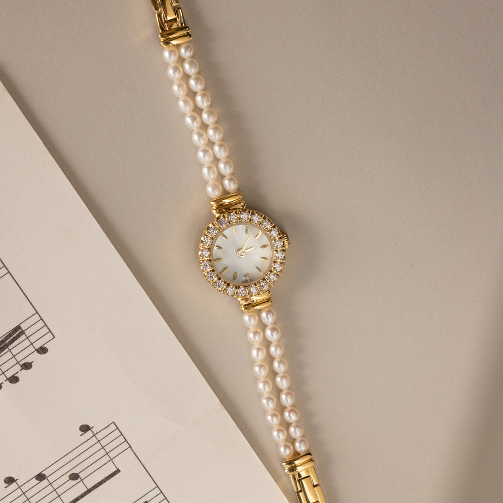 The Pearl Link Timepiece, featuring diamond accents and a jeweled face, sits beside sheet music on a beige surface, exuding timeless vintage elegance.