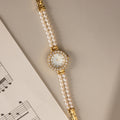 The Pearl Link Timepiece, featuring diamond accents and a jeweled face, sits beside sheet music on a beige surface, exuding timeless vintage elegance.