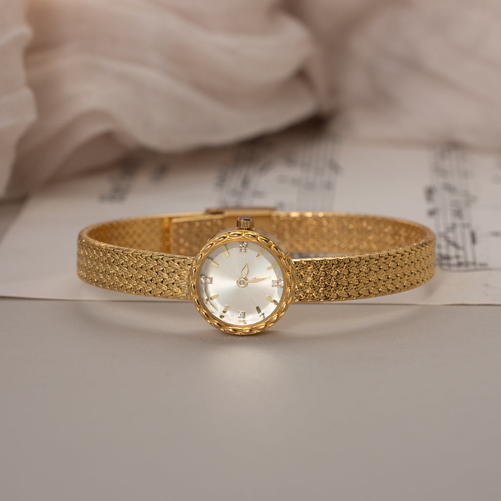 Side view of the round braided gold watch placed on sheet music with soft fabric in the background.