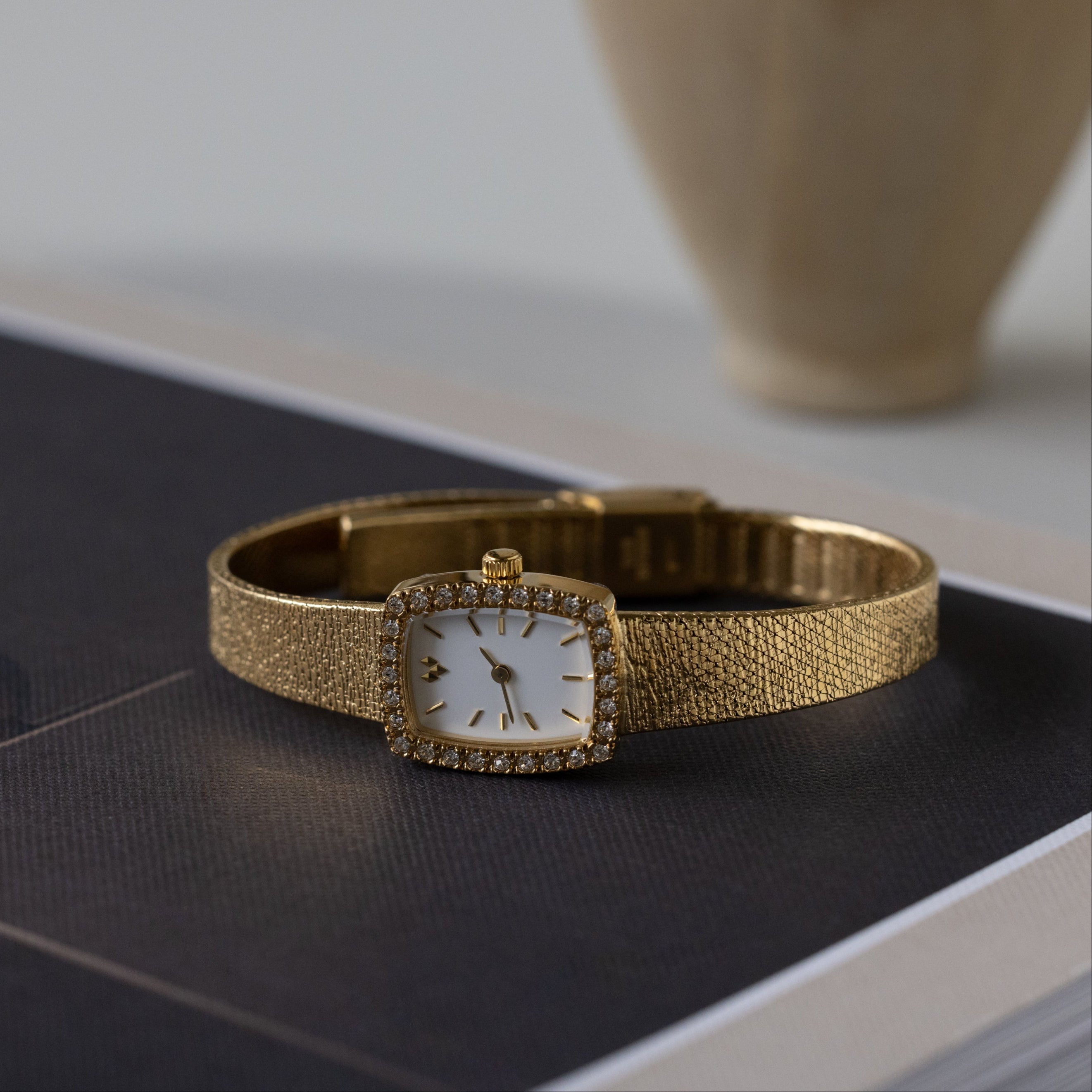 The Petite Mesh Timepiece, featuring a gold mesh band and a square diamond-studded face, rests elegantly atop a closed black book.