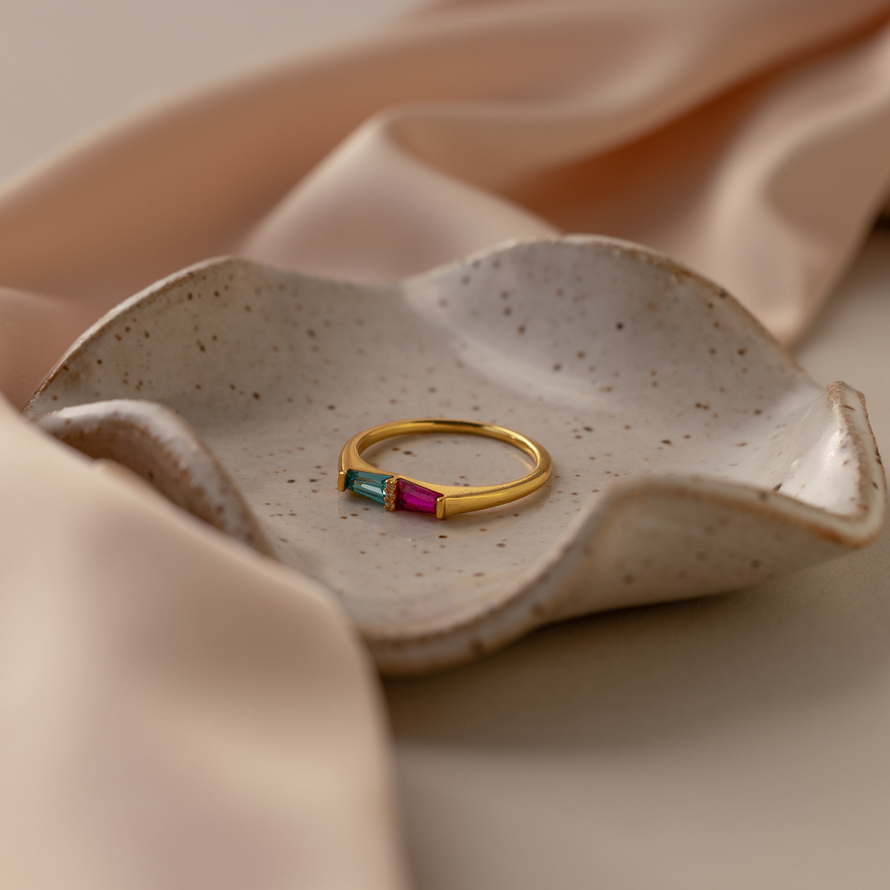 The Toi et Moi Tapered Birthstone Ring, featuring blue and pink baguette-cut stones, rests on a speckled ceramic dish with beige satin fabric in the background.