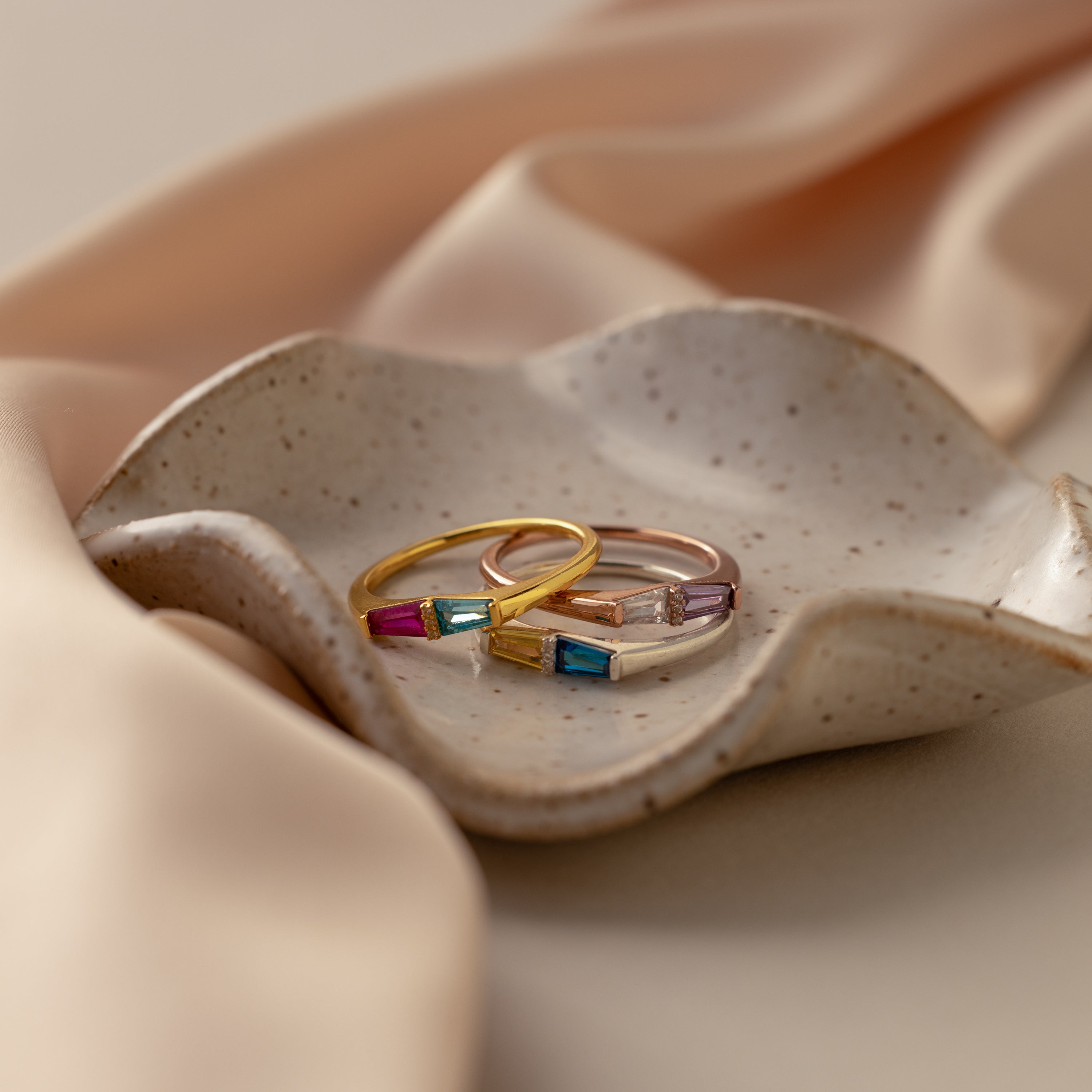 Three Toi et Moi Tapered Birthstone Rings with colorful baguette-cut stones rest on a speckled ceramic dish atop beige satin fabric.