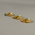 Four gold rings with colorful gemstones, including a Toi et Moi Birthstone Signet Ring, are arranged in a diagonal line on a neutral surface.