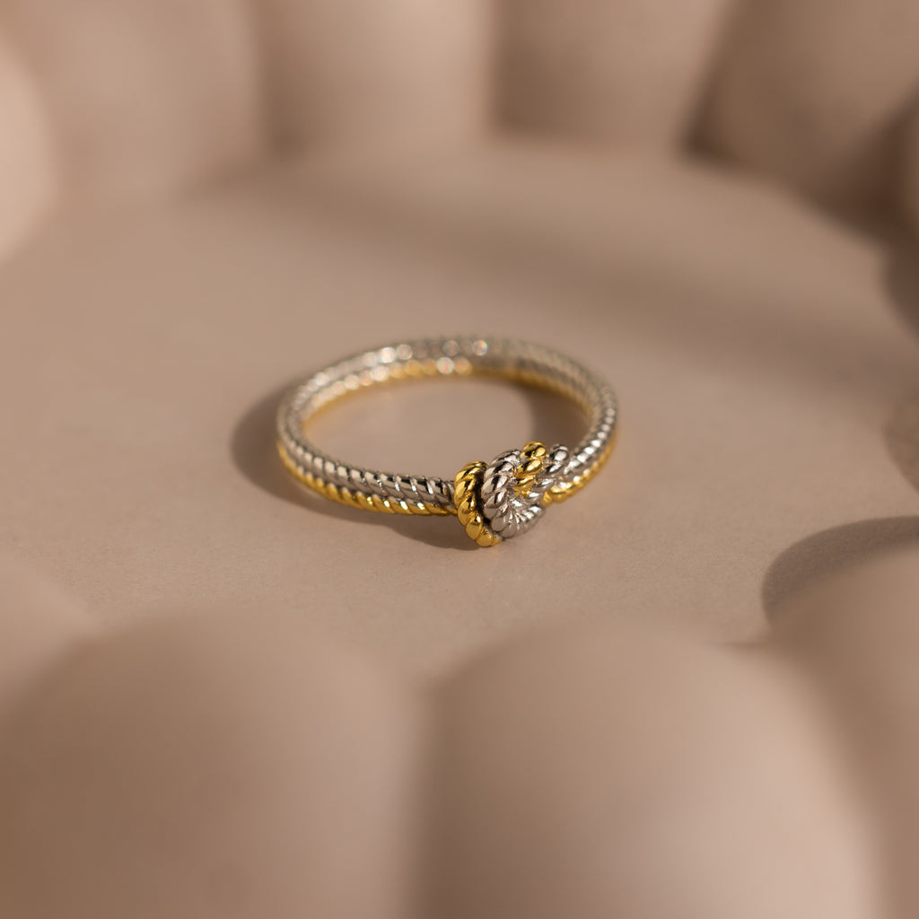 The Mixed Metal Knot Ring, featuring gold and silver in a twisted rope design with a knot detail, is displayed on a soft beige surface—making it a meaningful relationship gift.