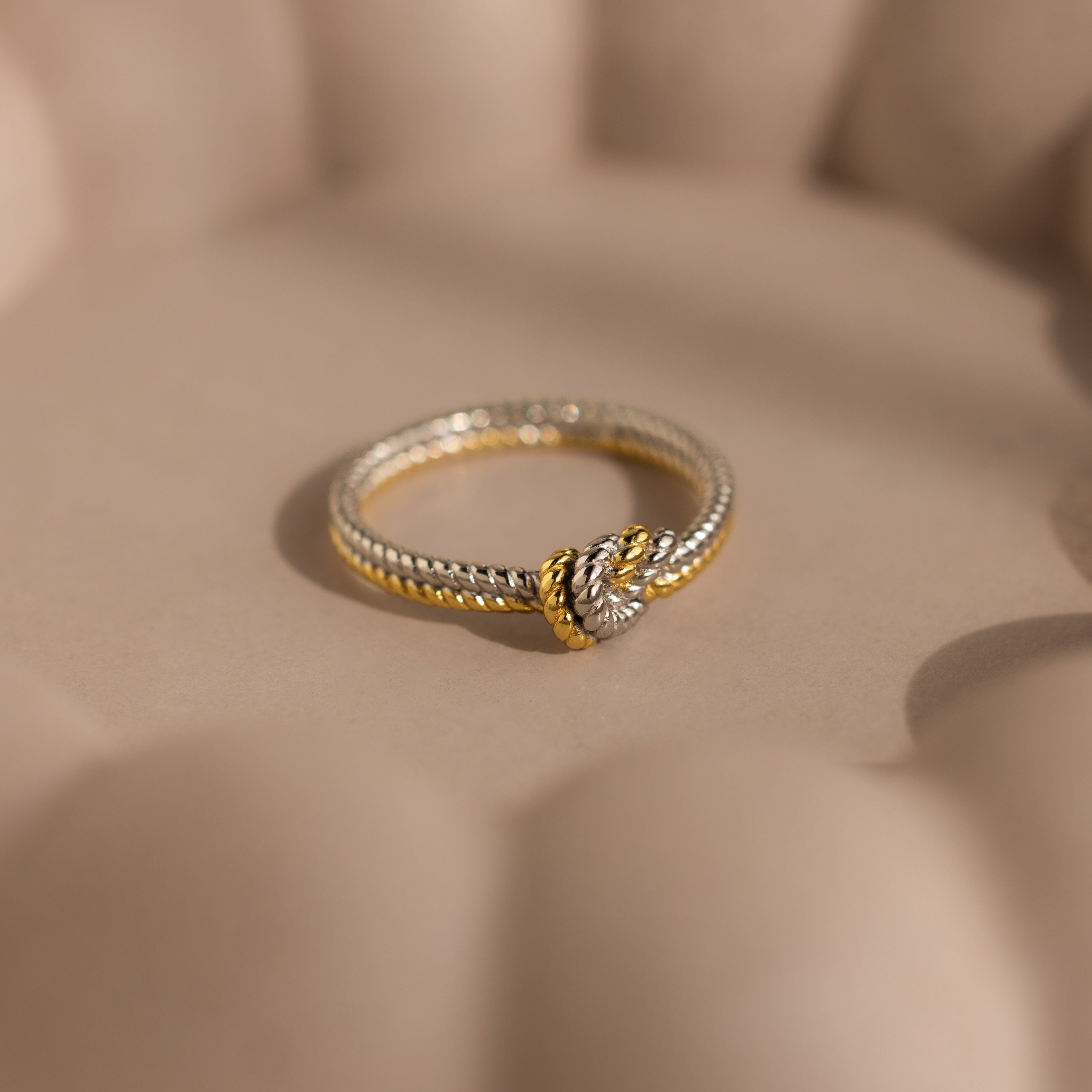 The Mixed Metal Knot Ring, featuring gold and silver in a twisted rope design with a knot detail, is displayed on a soft beige surface—making it a meaningful relationship gift.