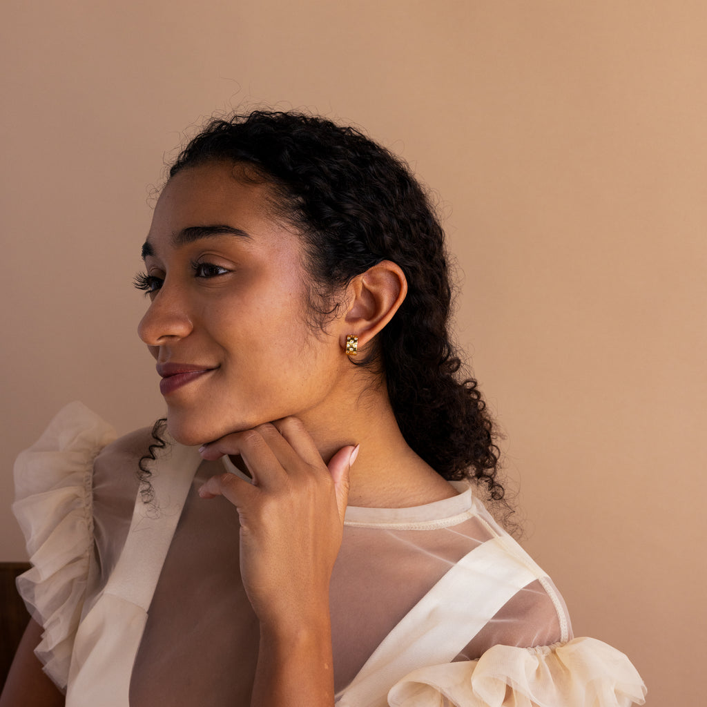 Woman with curly hair and gold earrings, smiling and resting her chin on her hand, wearing a sheer cream top and stylish Pave Checkered Huggies.