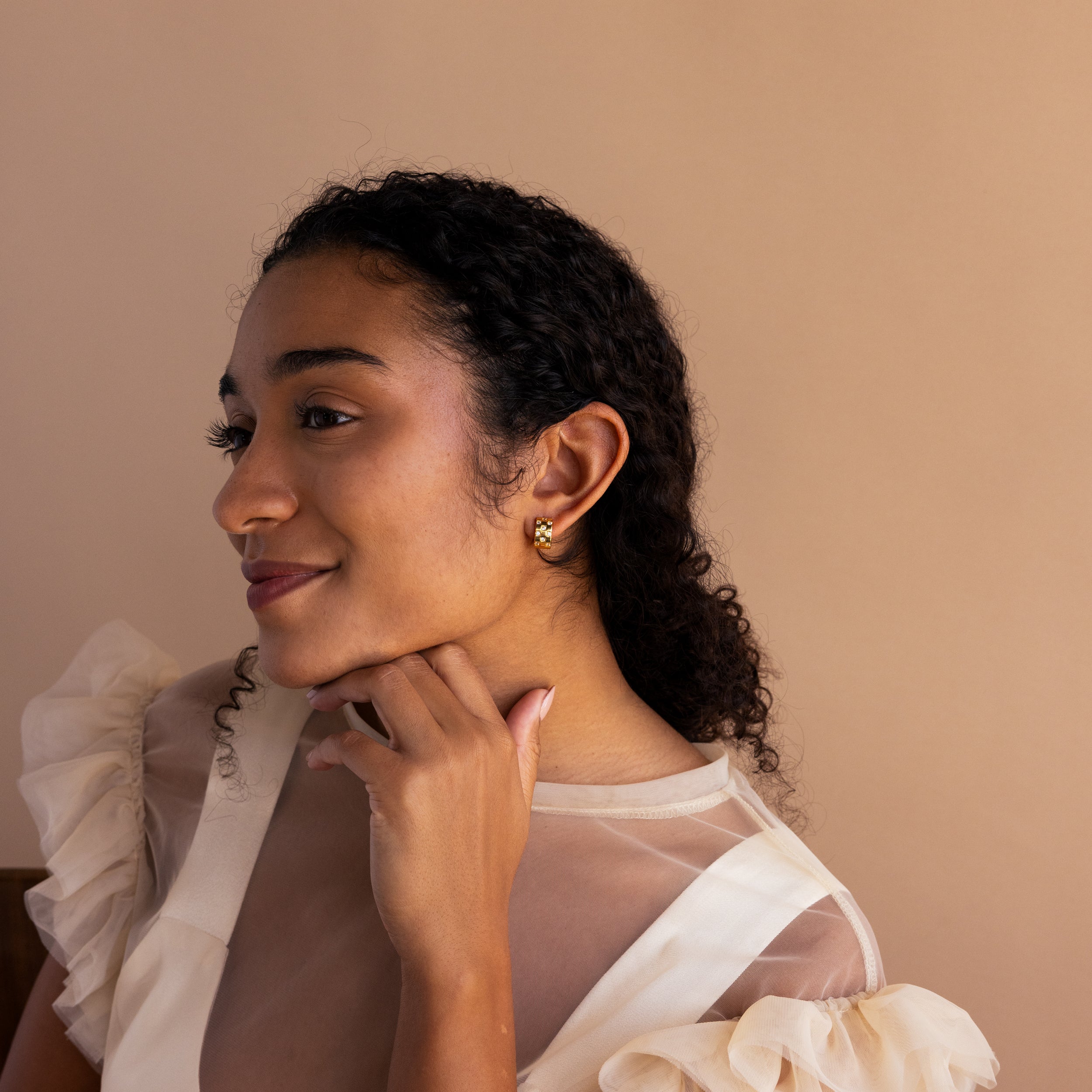 Woman with curly hair and gold earrings, smiling and resting her chin on her hand, wearing a sheer cream top and stylish Pave Checkered Huggies.