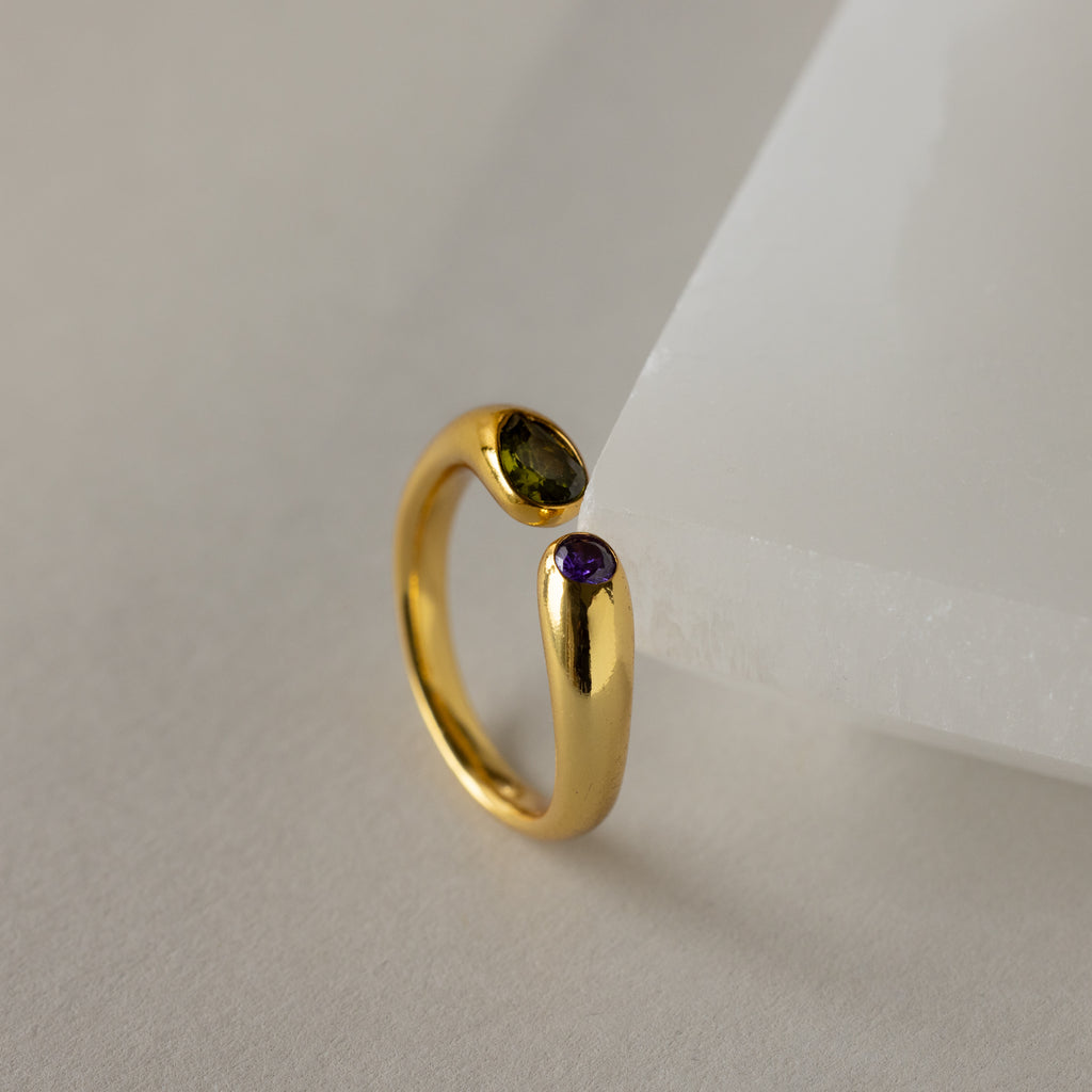 The Toi et Moi Birthstone Signet Ring in gold features a purple and a green gemstone, displayed on an angled white surface.