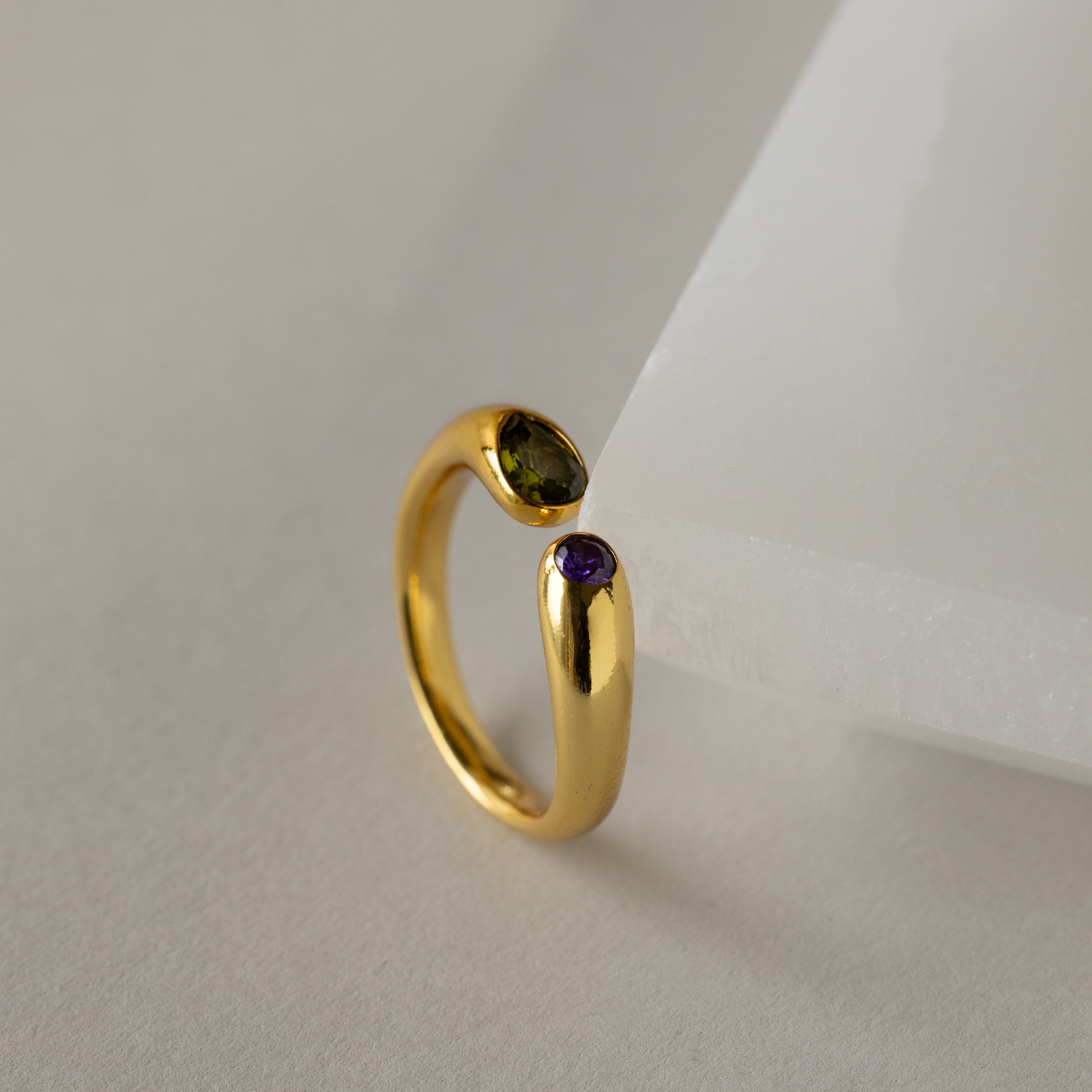 The Toi et Moi Birthstone Signet Ring in gold features a purple and a green gemstone, displayed on an angled white surface.