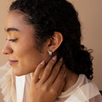 Woman with curly hair wearing Pave Checkered Huggies, a sheer top, and smiling softly as she touches her neck.
