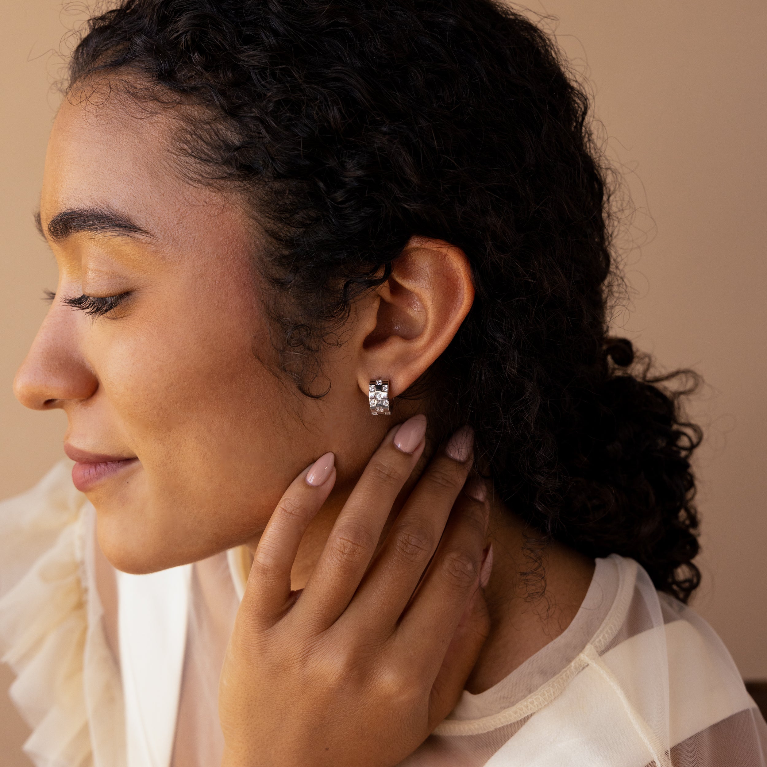 Woman with curly hair wearing Pave Checkered Huggies, a sheer top, and smiling softly as she touches her neck.