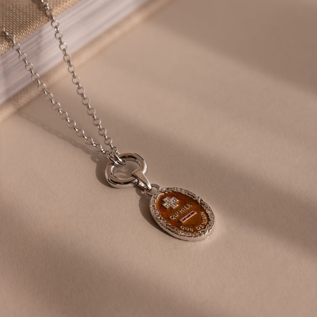 The Love Poem Pendant Necklace, featuring a silver chain and oval brown pendant, rests on a beige surface beside a book—a subtle token of love.