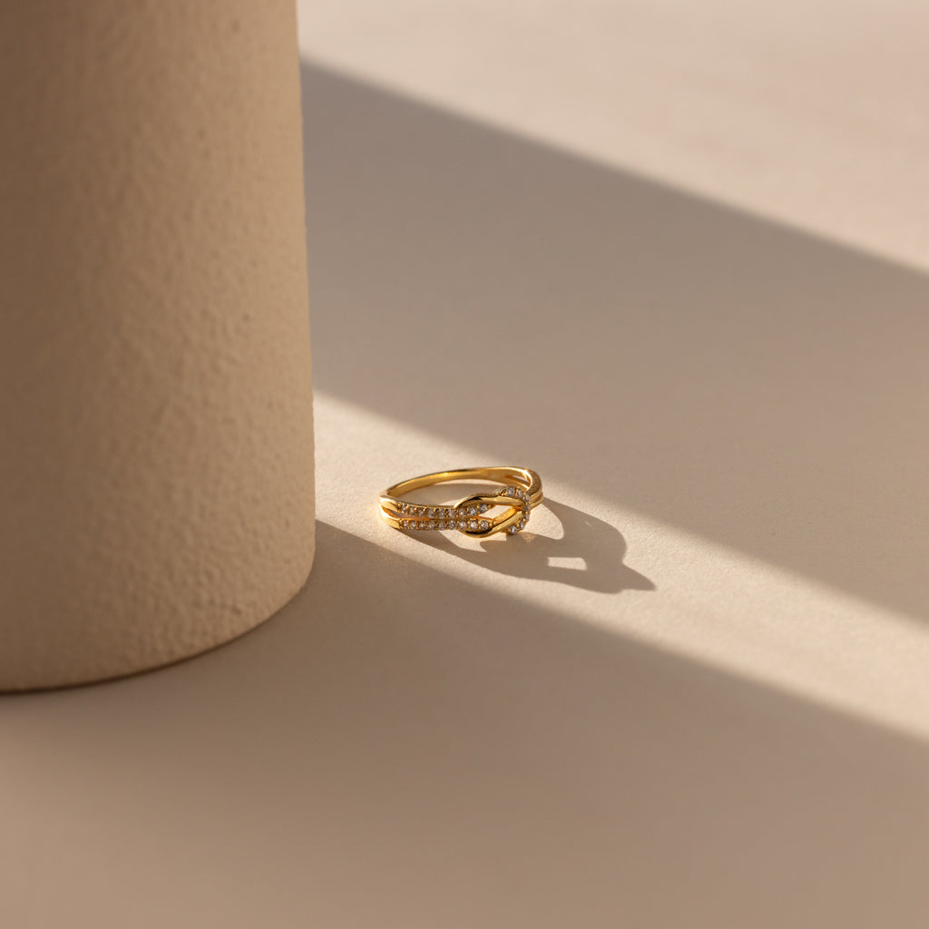 The Pave Double Knot Ring, featuring a gold pavé twist design, sparkles in the sunlight next to a textured cylindrical beige object.