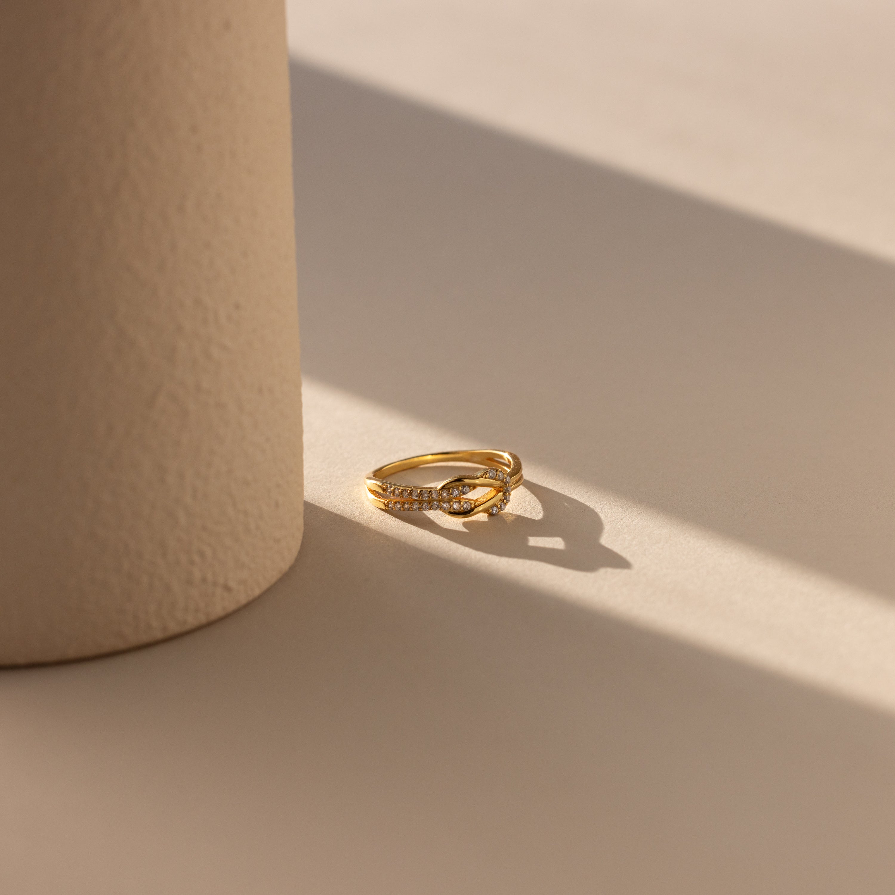 The Pave Double Knot Ring, featuring a gold pavé twist design, sparkles in the sunlight next to a textured cylindrical beige object.