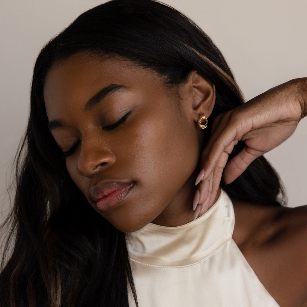 A woman with long dark hair exudes quiet luxury, eyes closed and hand near her face, wearing elegant gold hoops and Micro Triple Knot Earrings.