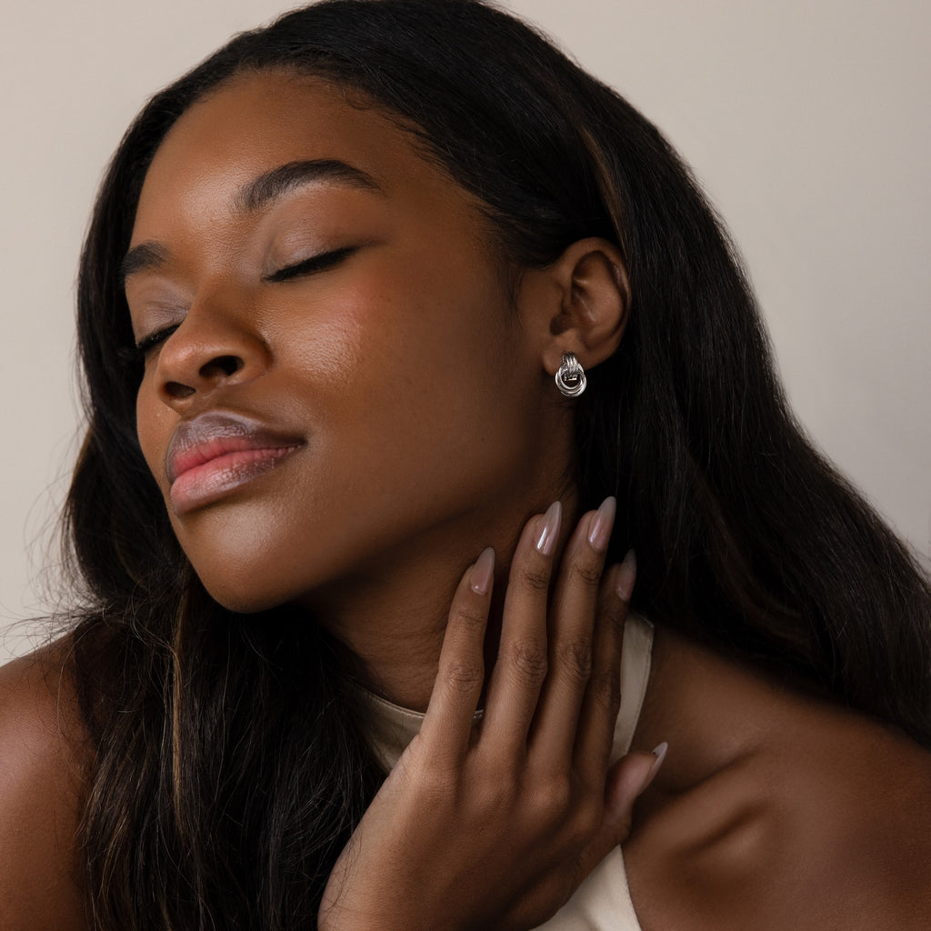 Woman with long dark hair smiles softly, eyes closed, touching her neck while wearing Micro Triple Knot Earrings and nude nail polish.