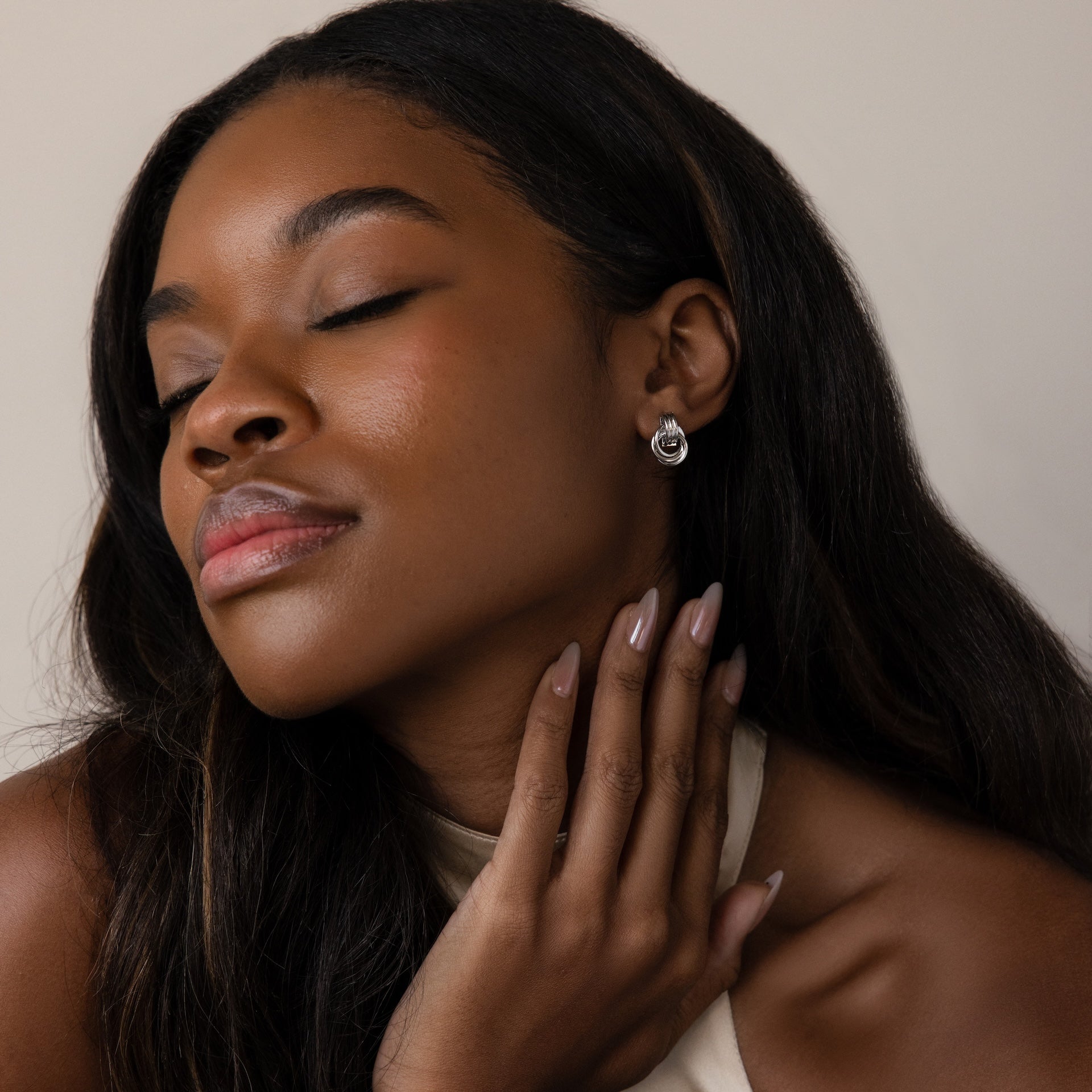 Woman with long dark hair smiles softly, eyes closed, touching her neck while wearing Micro Triple Knot Earrings and nude nail polish.