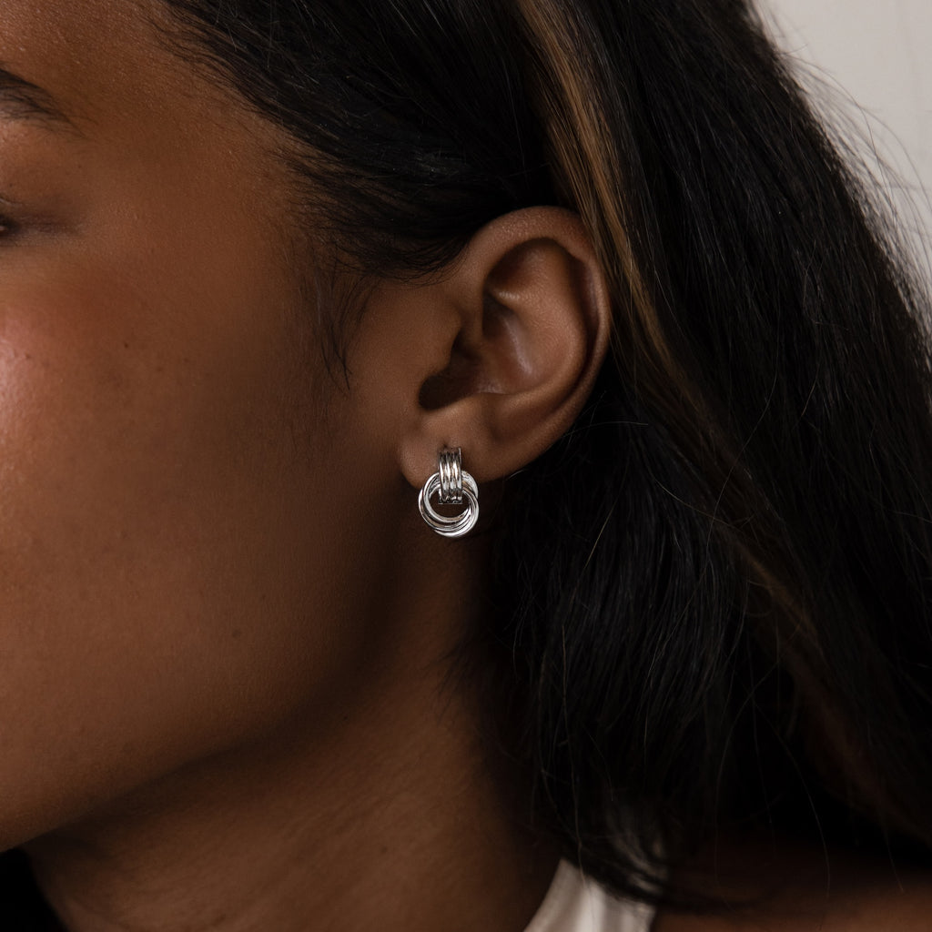 Close-up of a person with long dark hair and brown skin wearing Micro Triple Knot Earrings in Silver, showcasing quiet luxury style.