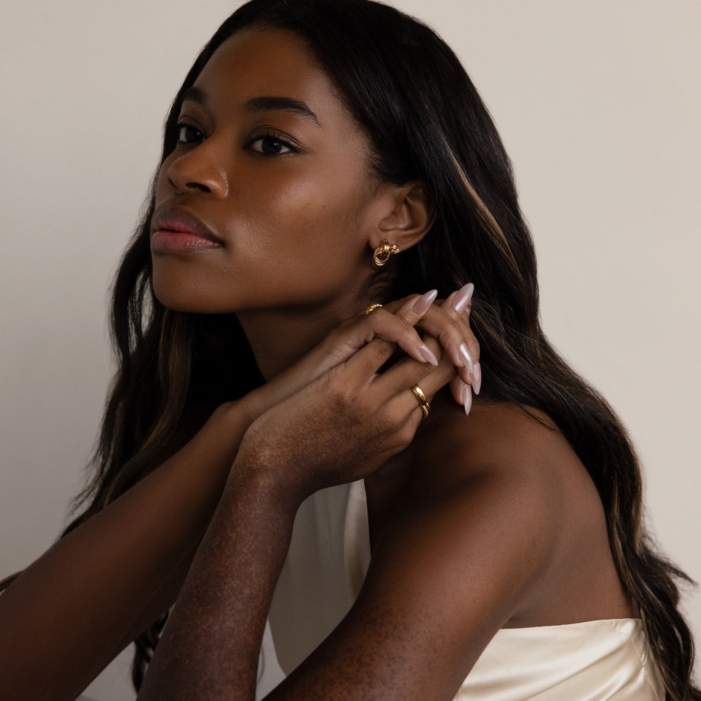 A woman with long dark hair and gold Micro Triple Knot Earrings poses against a neutral background in a cream top, radiating quiet luxury.