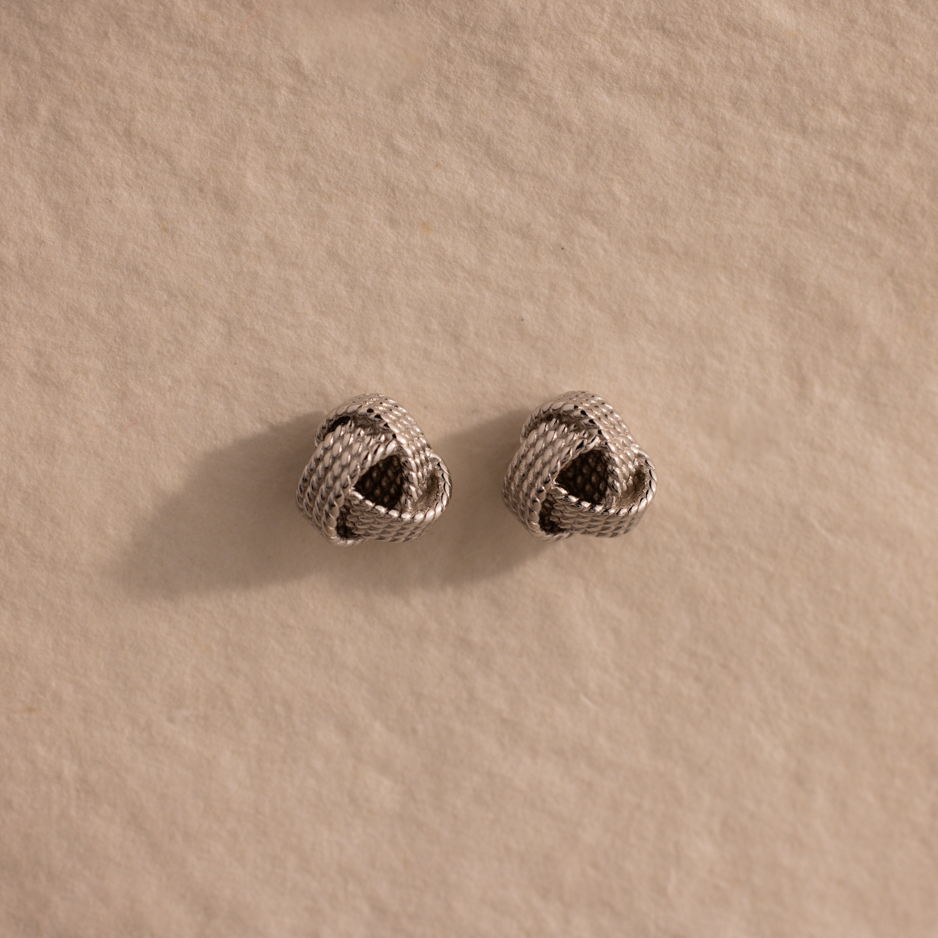 The Rope Knot Studs in Silver, with their elegant knot design, rest on a textured beige surface—timeless accessories that are perfect for any jewelry collection.