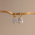 Celestial Baroque Pearl Charm Hoops in 18K Gold feature irregular white pearls suspended from gold hoops, softly gleaming against a neutral beige backdrop.