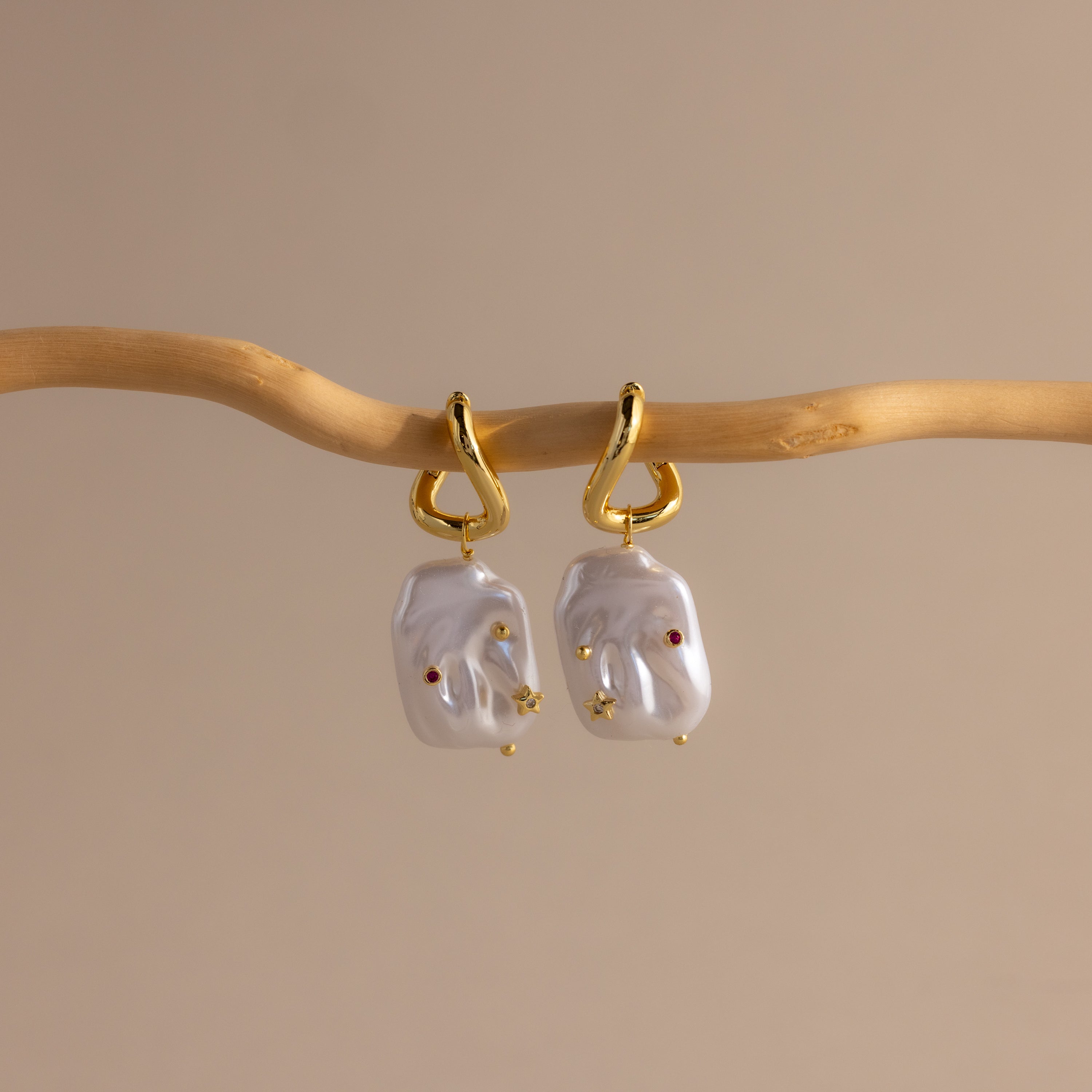 Celestial Baroque Pearl Charm Hoops in 18K Gold feature irregular white pearls suspended from gold hoops, softly gleaming against a neutral beige backdrop.