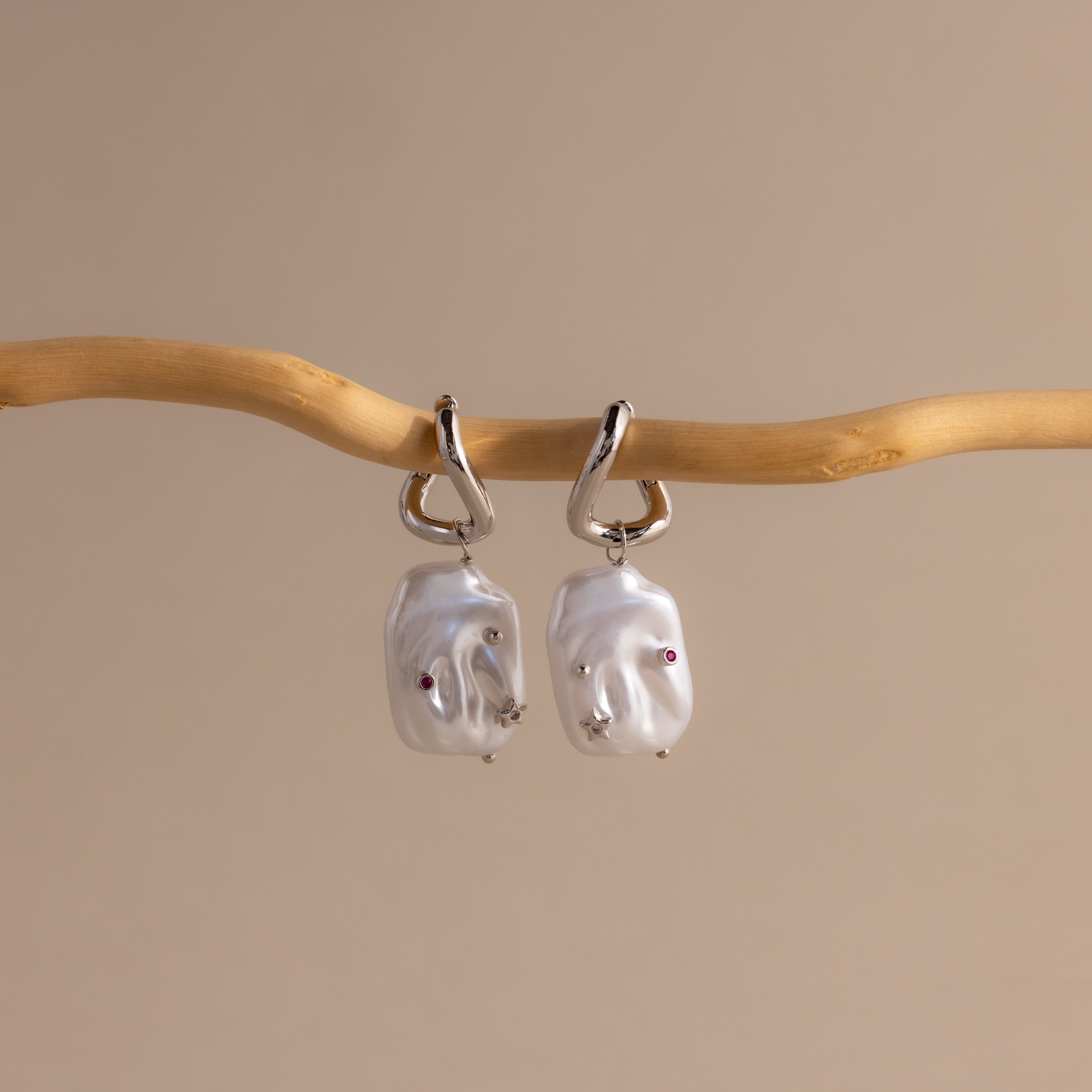 The Celestial Baroque Pearl Charm Hoops in White Gold feature two irregular silver pearls that dangle elegantly from hoops, displayed against a beige backdrop for a subtle yet stunning effect.