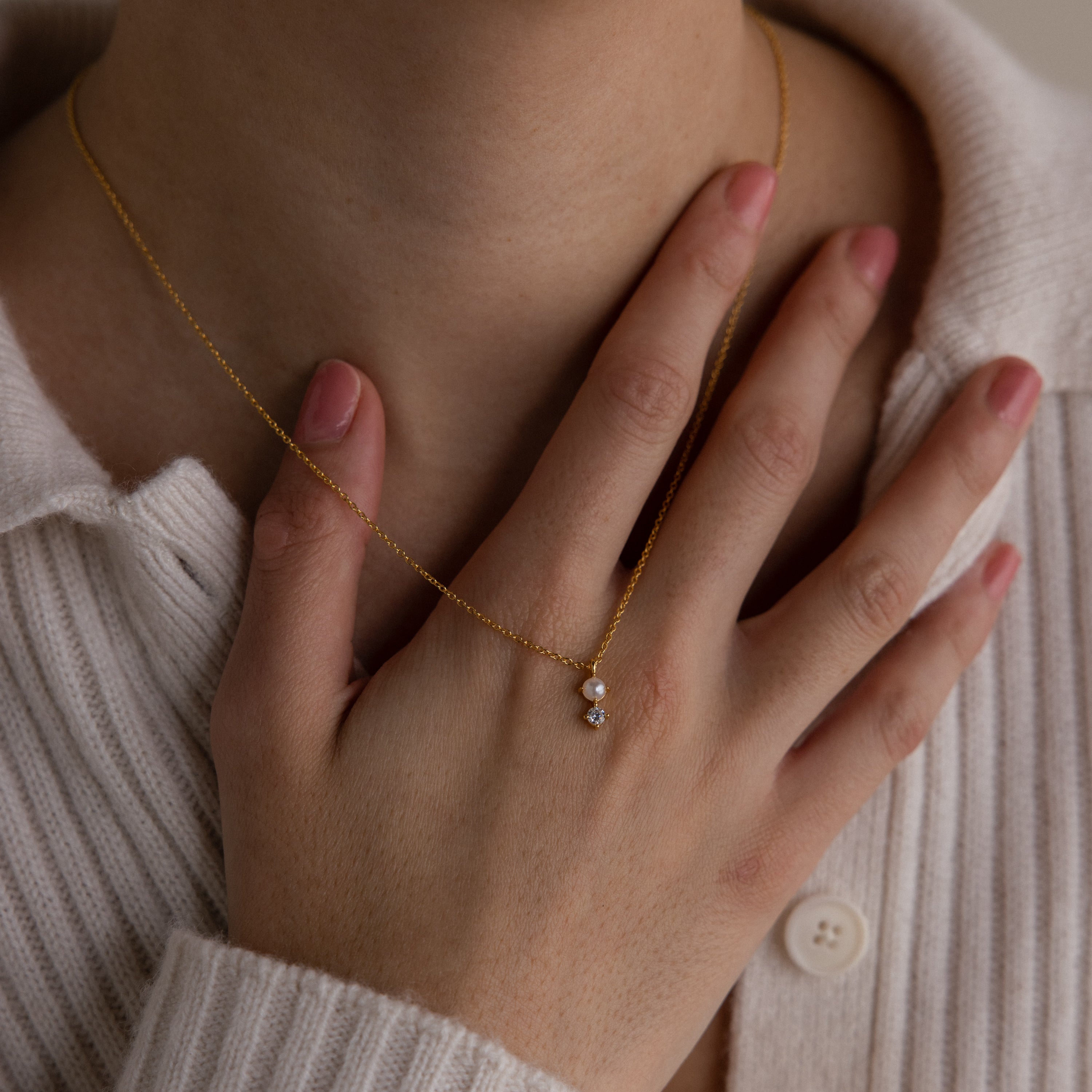 Wearing a cream sweater, a person touches the Pearl Diamond Drop Necklace, which features a delicate small pendant on their neck.