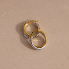 The Twisted Mixed Metal Hoops in gold and silver are displayed on a beige, lightly textured surface, highlighting their modern chic design.