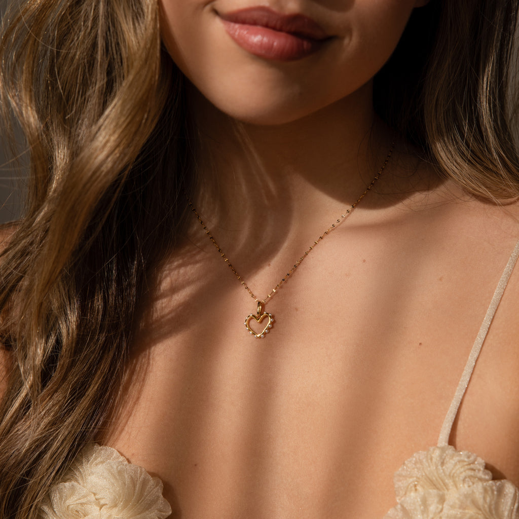 A woman wears a cream floral top and an 18K gold Classic Pearl Heart Charm necklace.