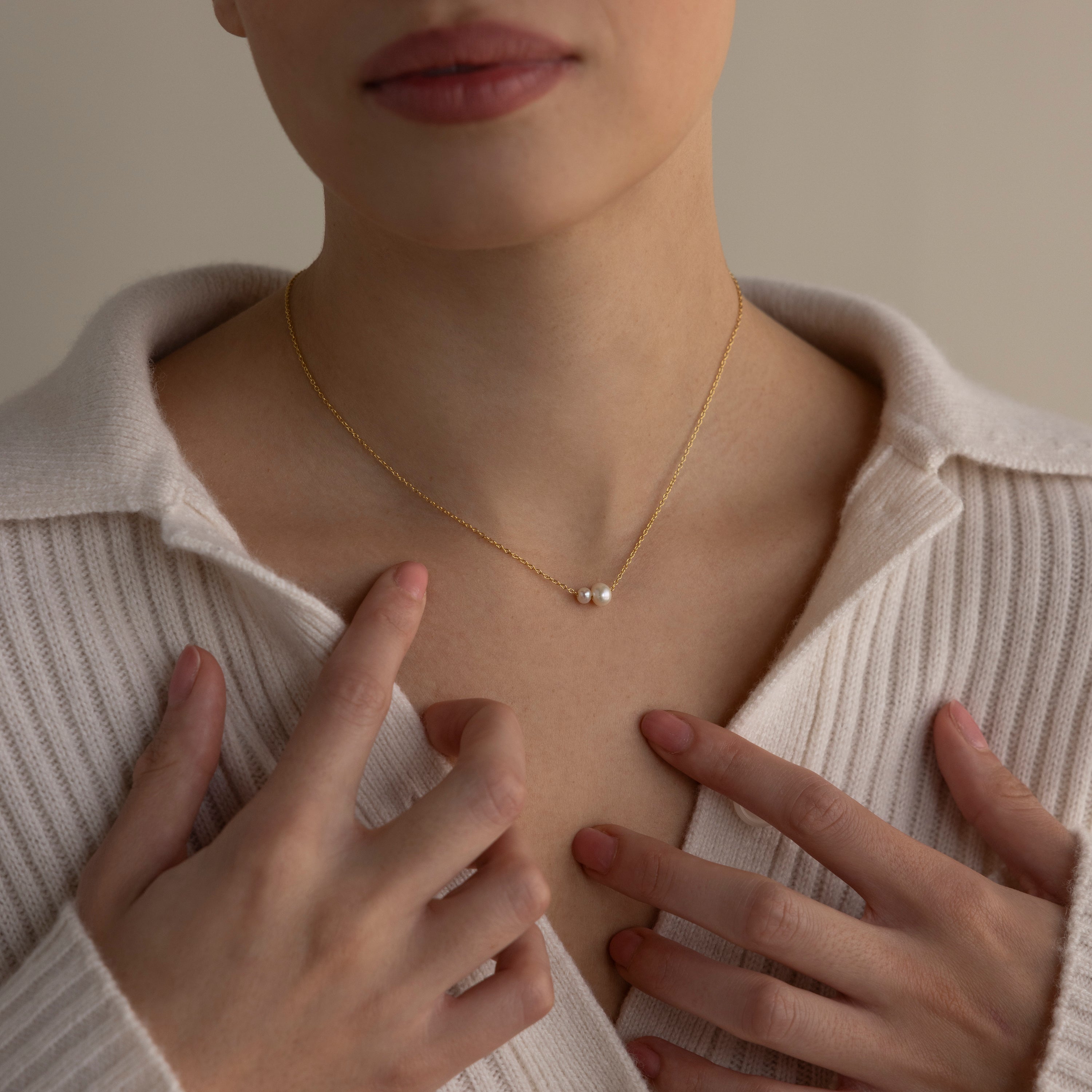 Woman wearing a cream ribbed shirt and the Dainty Duo Pearl Necklace, featuring two pearls, gently touching her chest.