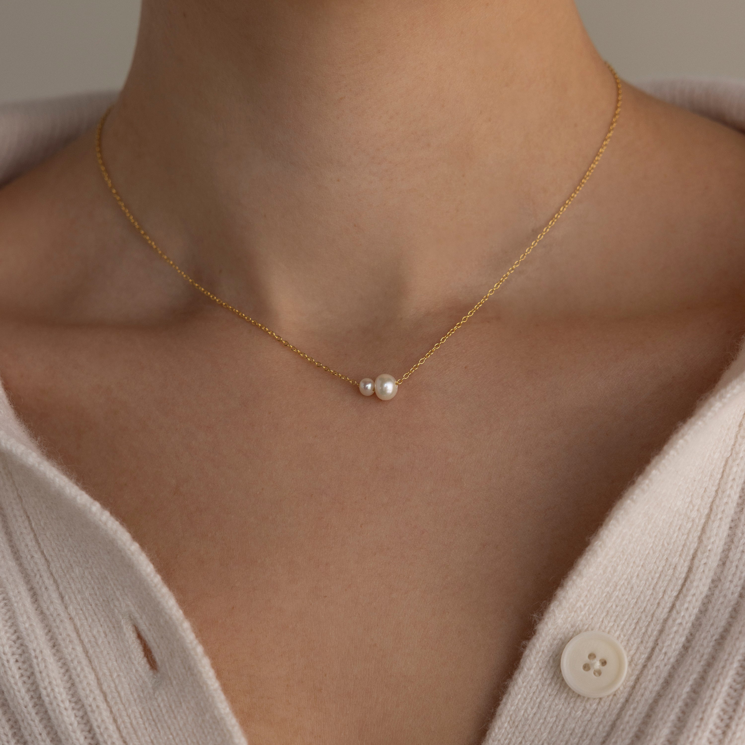 A close-up of a woman wearing the Dainty Duo Pearl Necklace, featuring two small pearls, styled with a cream sweater.