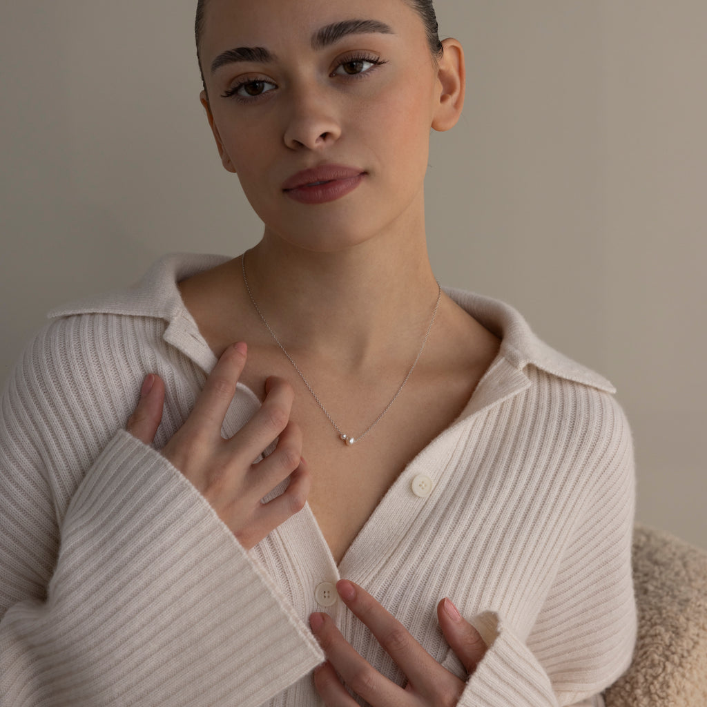 Woman in a cream ribbed sweater touches the Dainty Duo Pearl Necklace in Silver, gazing softly at the camera—a perfect gift for loved ones.