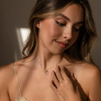A woman with long hair, wearing a beige top and smiling gently while looking down, showcases the Classic Pearl Heart Charm in Sterling Silver necklace.