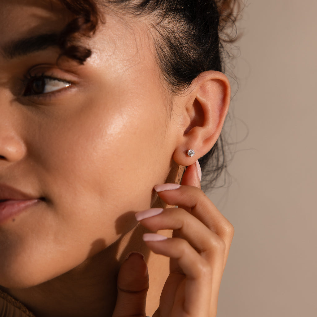Model touches a single small diamond stud earring in her earlobe with manicured fingers.