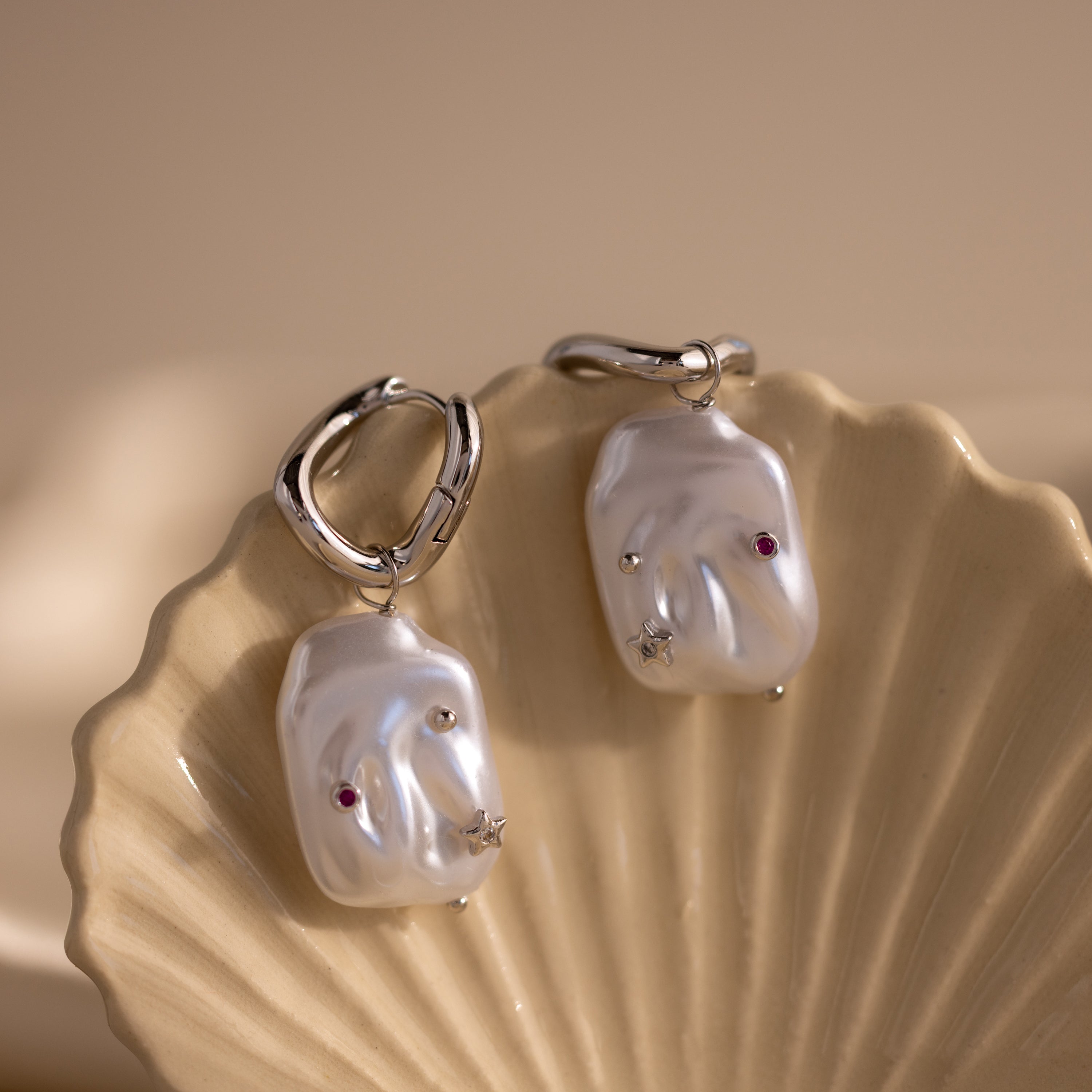 The Celestial Baroque Pearl Charm Hoops, featuring silver statement hoops with irregular pearl charms and small gems, are displayed on a shell dish for the perfect baroque look.
