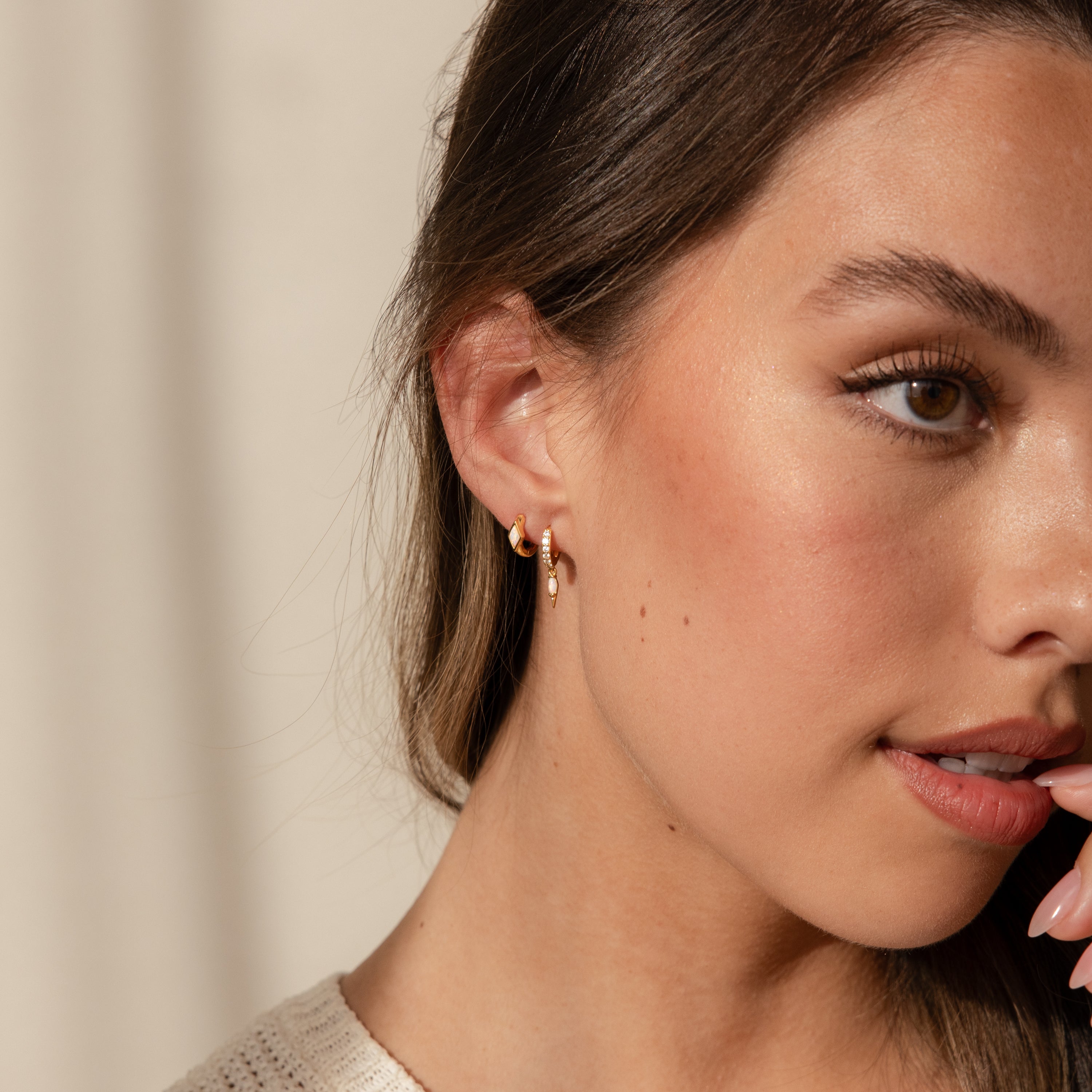 A woman with long brown hair wears Rhombus Topaz Huggies and a neutral top, gazing thoughtfully to the side.