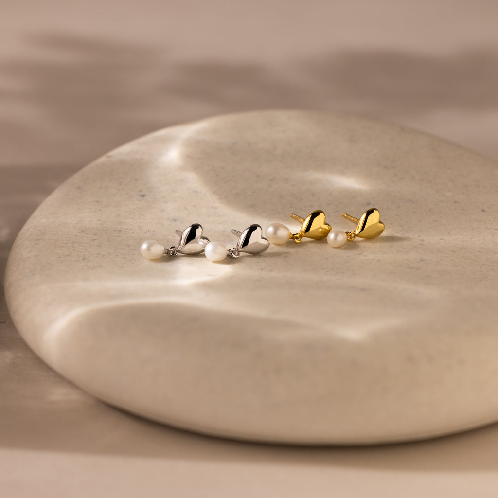 The Heart Pearl Drop Earrings feature heart-shaped studs and elegant pearl drops, shown on a smooth, round stone surface.
