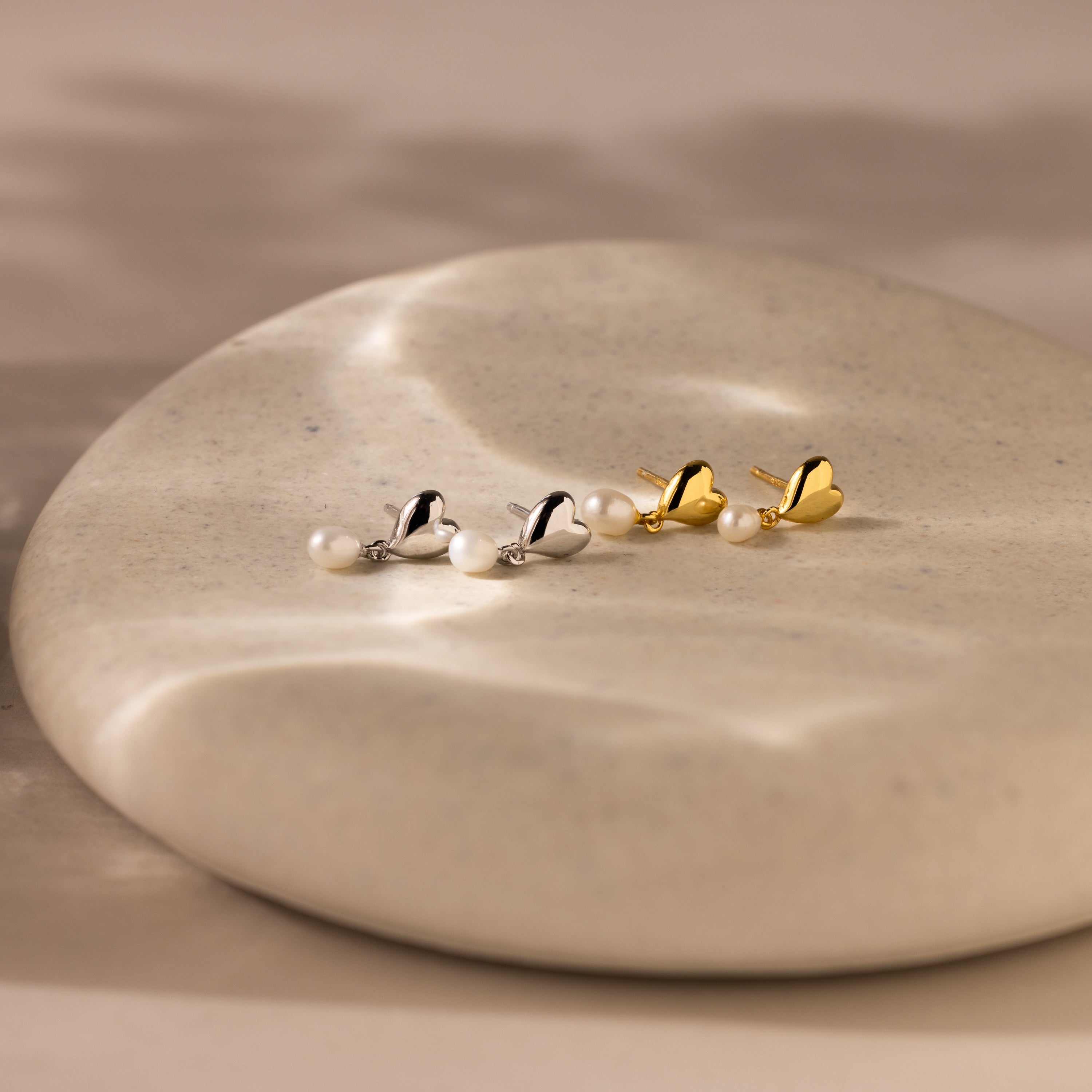 The Heart Pearl Drop Earrings feature heart-shaped studs and elegant pearl drops, shown on a smooth, round stone surface.