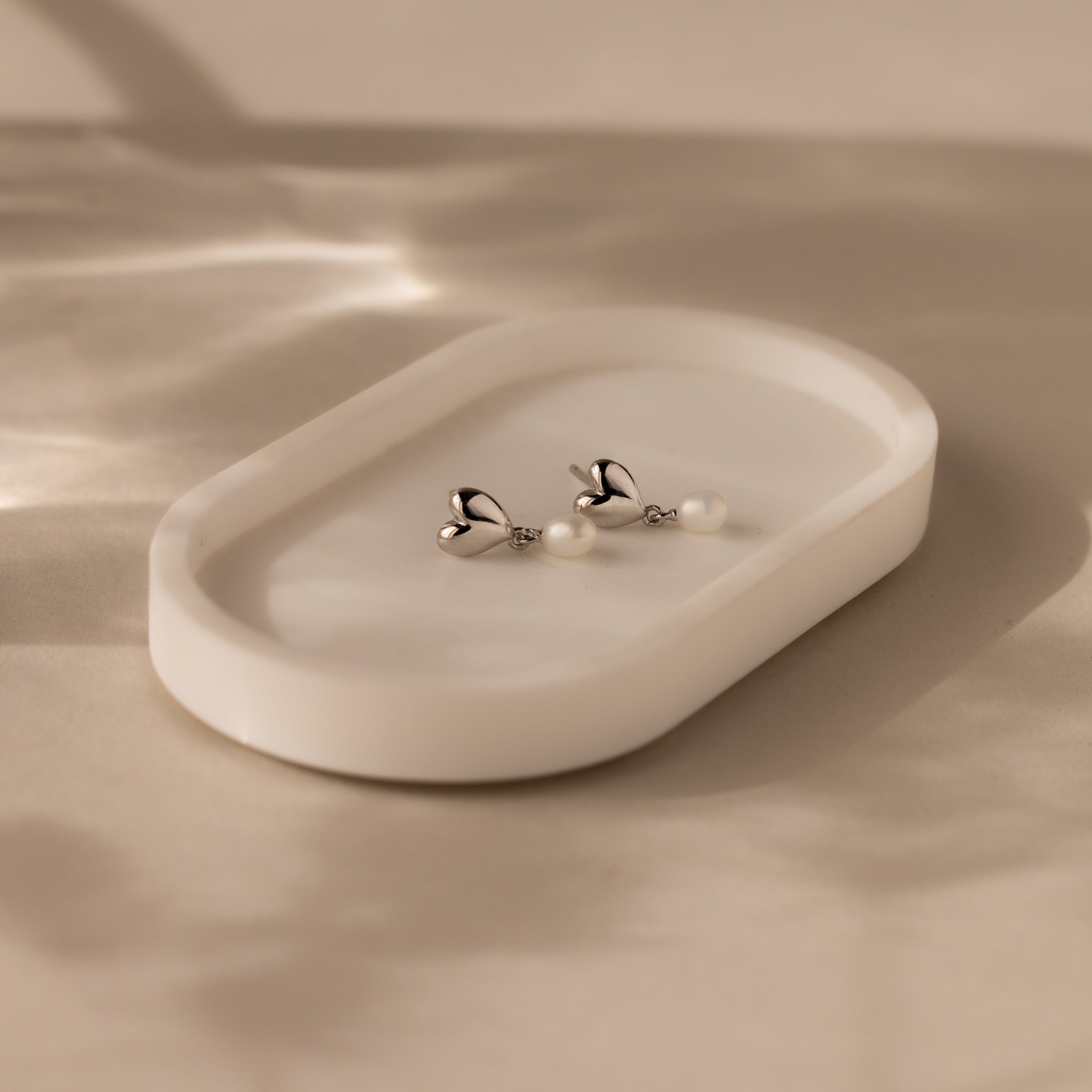 Heart Pearl Drop Earrings featuring delicate pearls in a heart shape rest on a white oval dish, softly lit by natural light—ideal for any occasion.