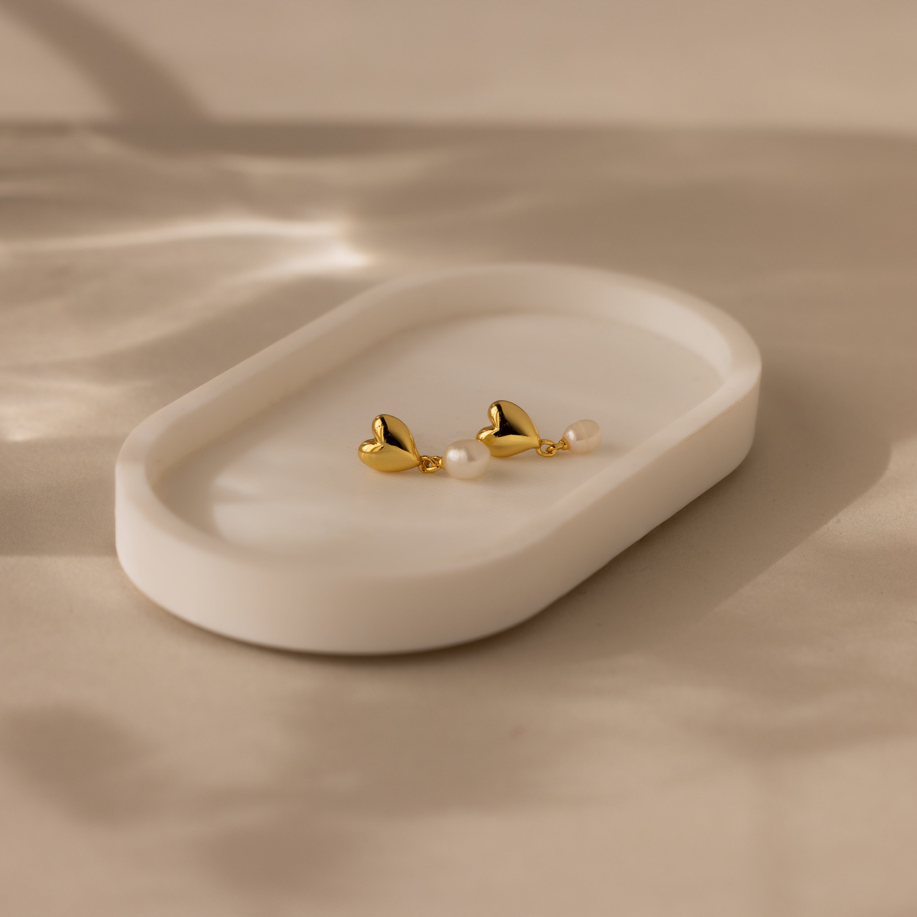 Heart Pearl Drop Earrings in 18K Gold with pearls are displayed on a white oval dish set against a soft beige background.