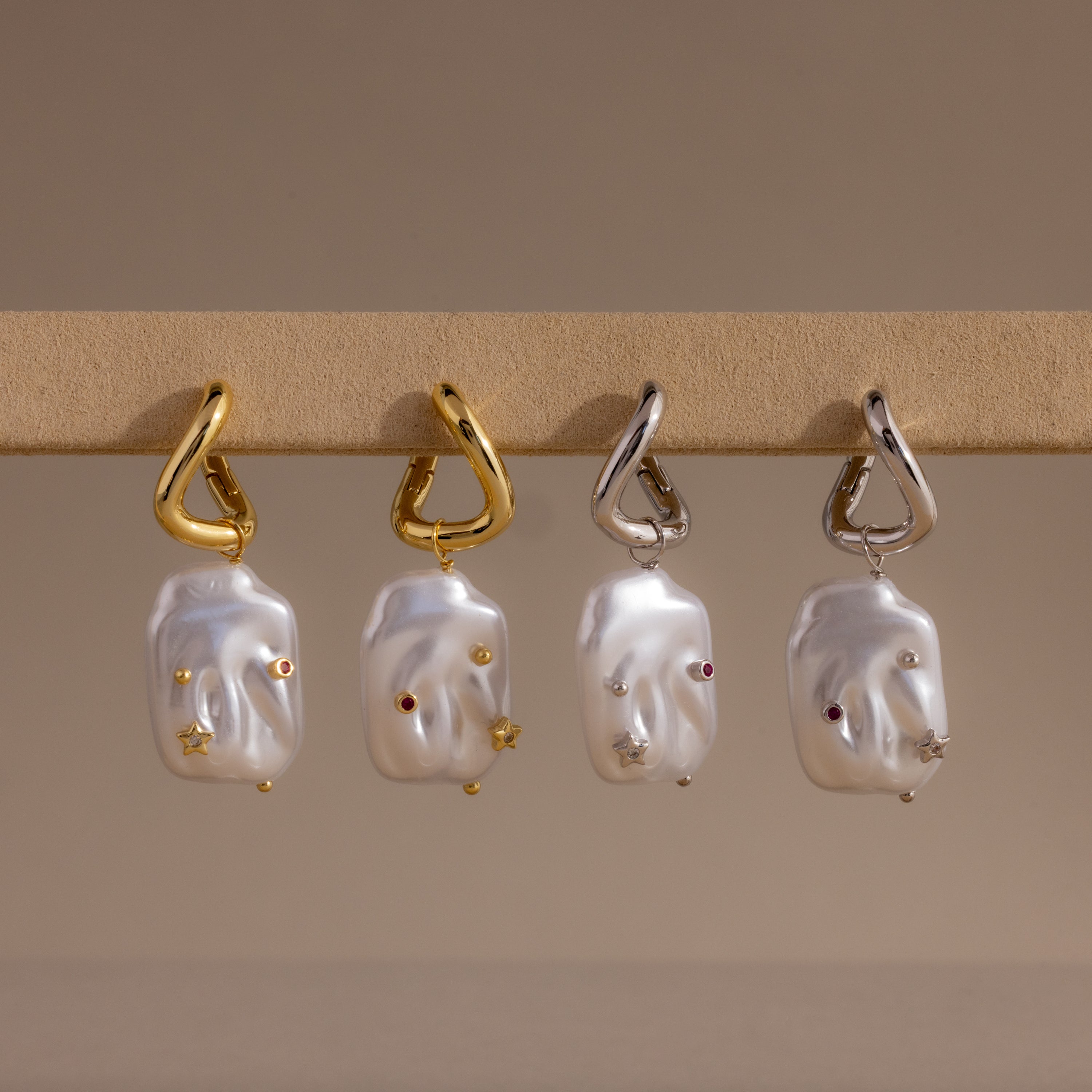 The Celestial Baroque Pearl Charm Hoops feature four baroque pearl earrings with metallic accents—two with gold hooks and two with silver—making elegant statement pieces displayed on a beige surface.