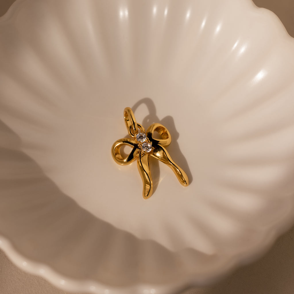 The Diamond Bow Charm is a gold, bow-shaped pendant with a small clear gemstone, displayed in a white scalloped dish—a charming coquette accessory perfect for any collection.