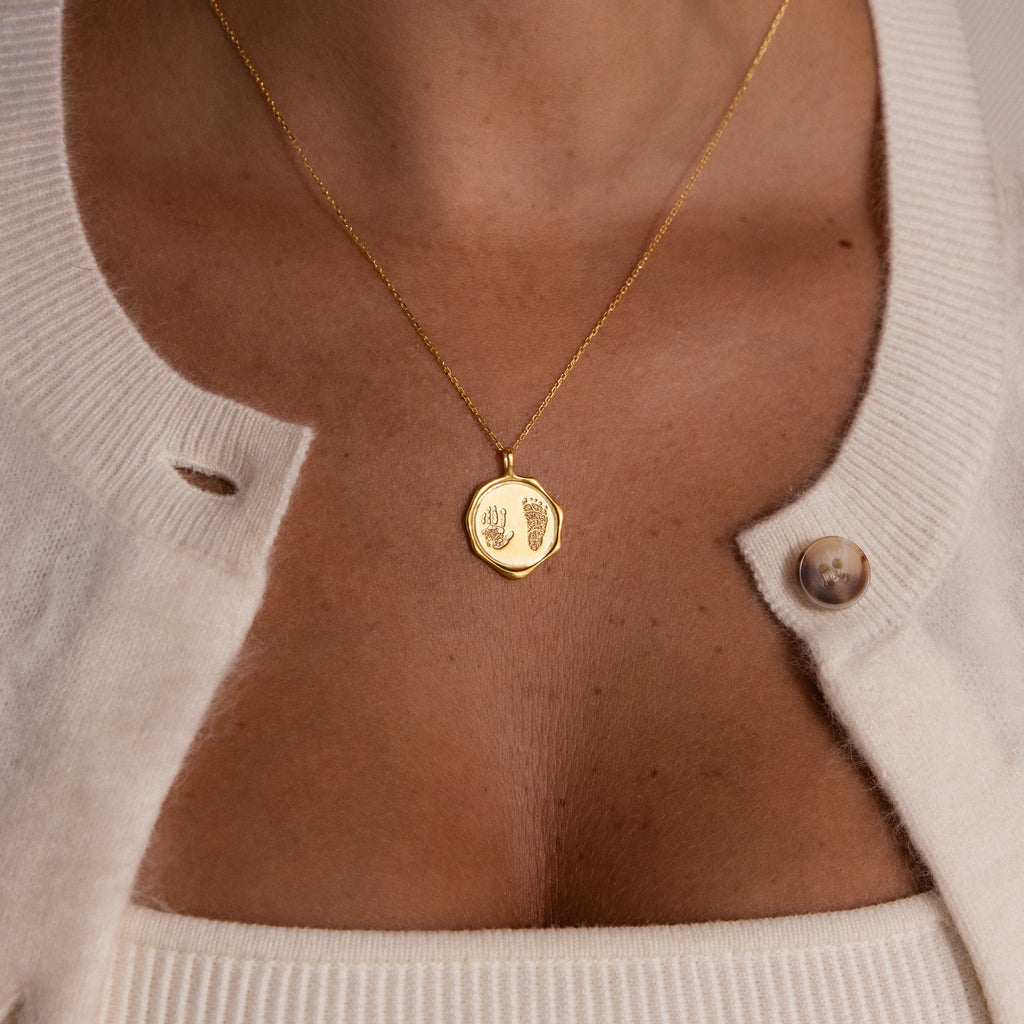 The Baby Hand & Footprint Necklace in 18K Gold, engraved with baby prints and styled with a cream cardigan and top, is a meaningful keepsake—ideal for fans of personalized baby jewelry.