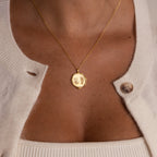 The Baby Hand & Footprint Necklace in 18K Gold, engraved with baby prints and styled with a cream cardigan and top, is a meaningful keepsake—ideal for fans of personalized baby jewelry.