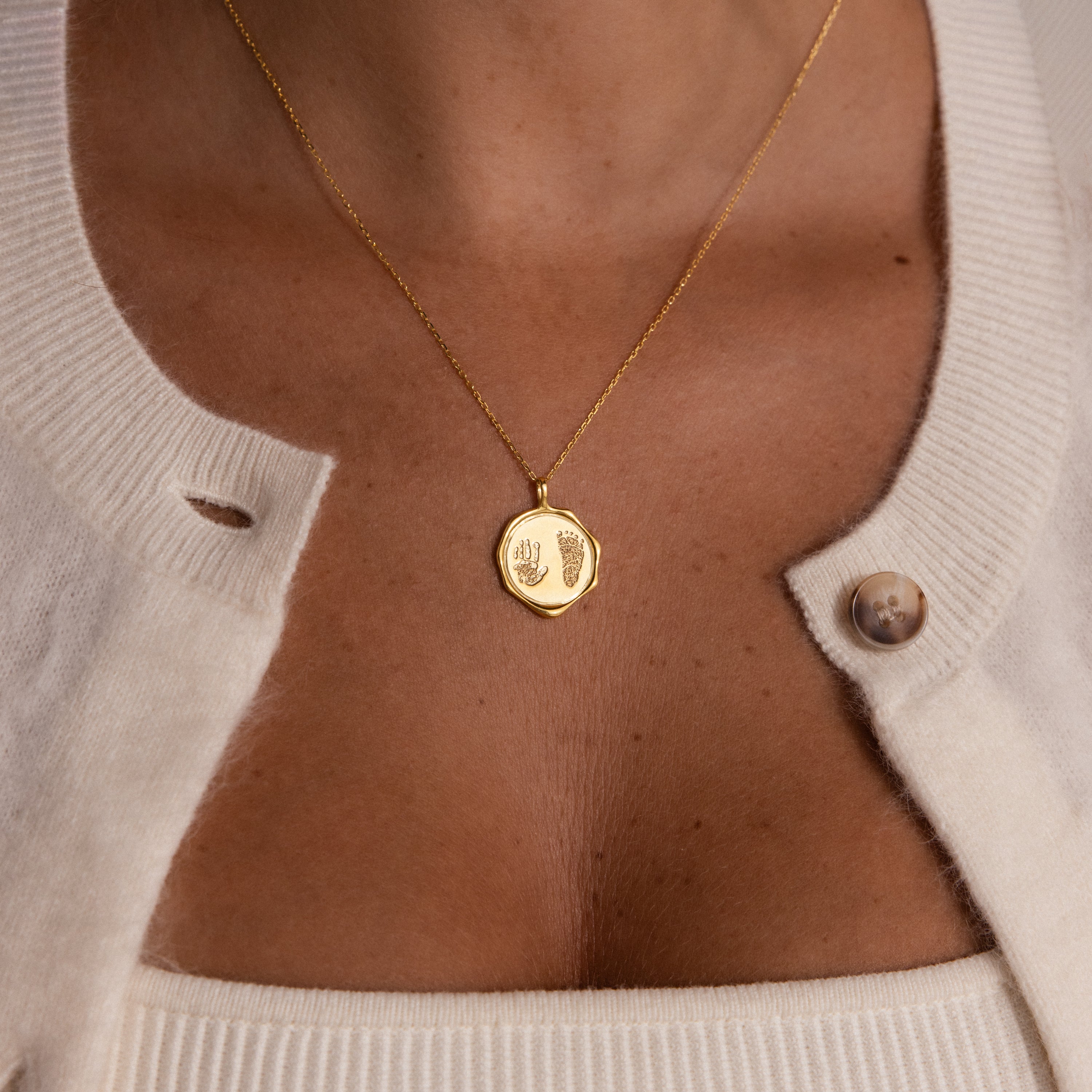The Baby Hand & Footprint Necklace in 18K Gold, engraved with baby prints and styled with a cream cardigan and top, is a meaningful keepsake—ideal for fans of personalized baby jewelry.